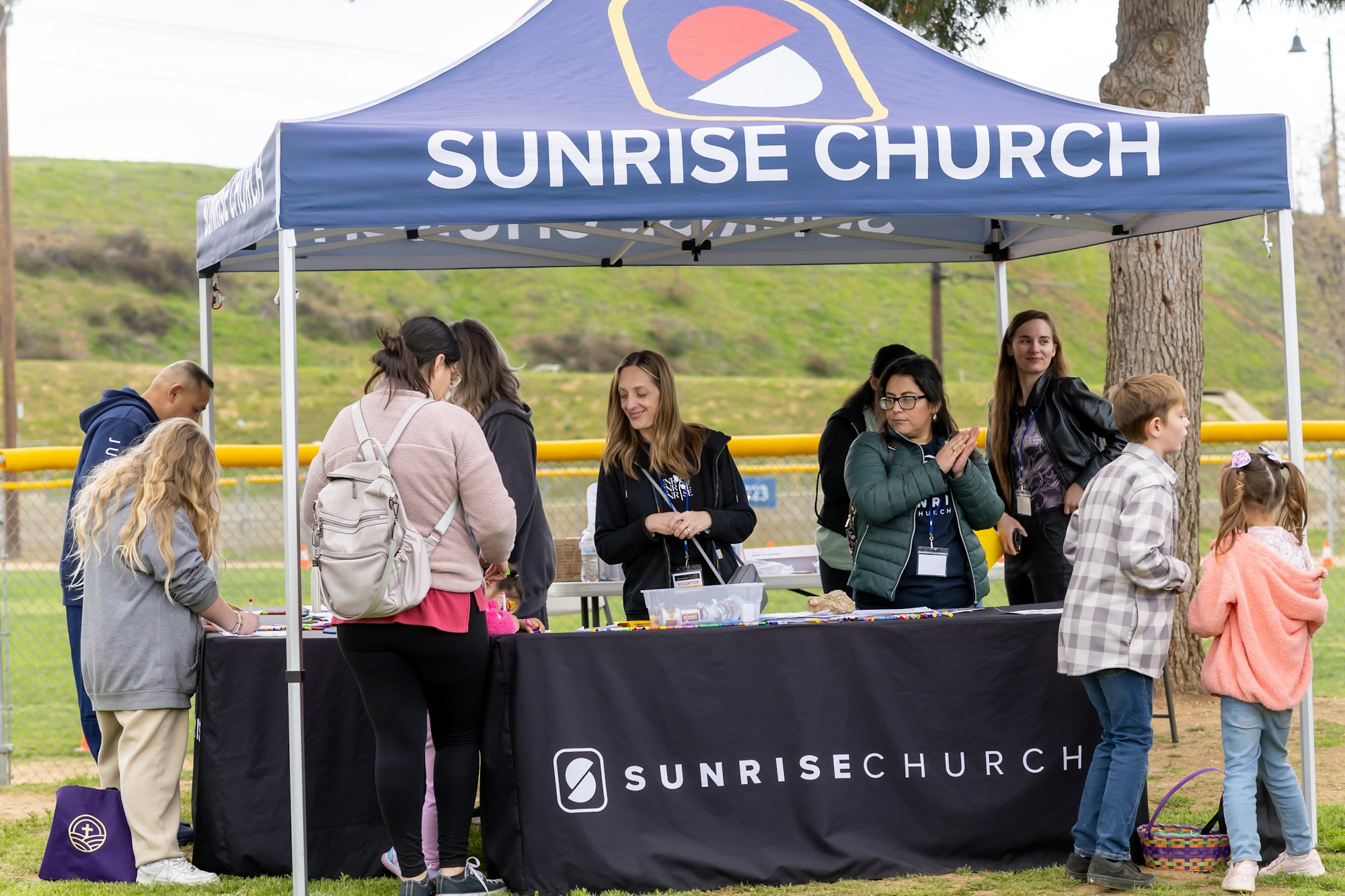 Outdoor church event with people gathered around a "Sunrise Church" booth under a canopy in a park setting.