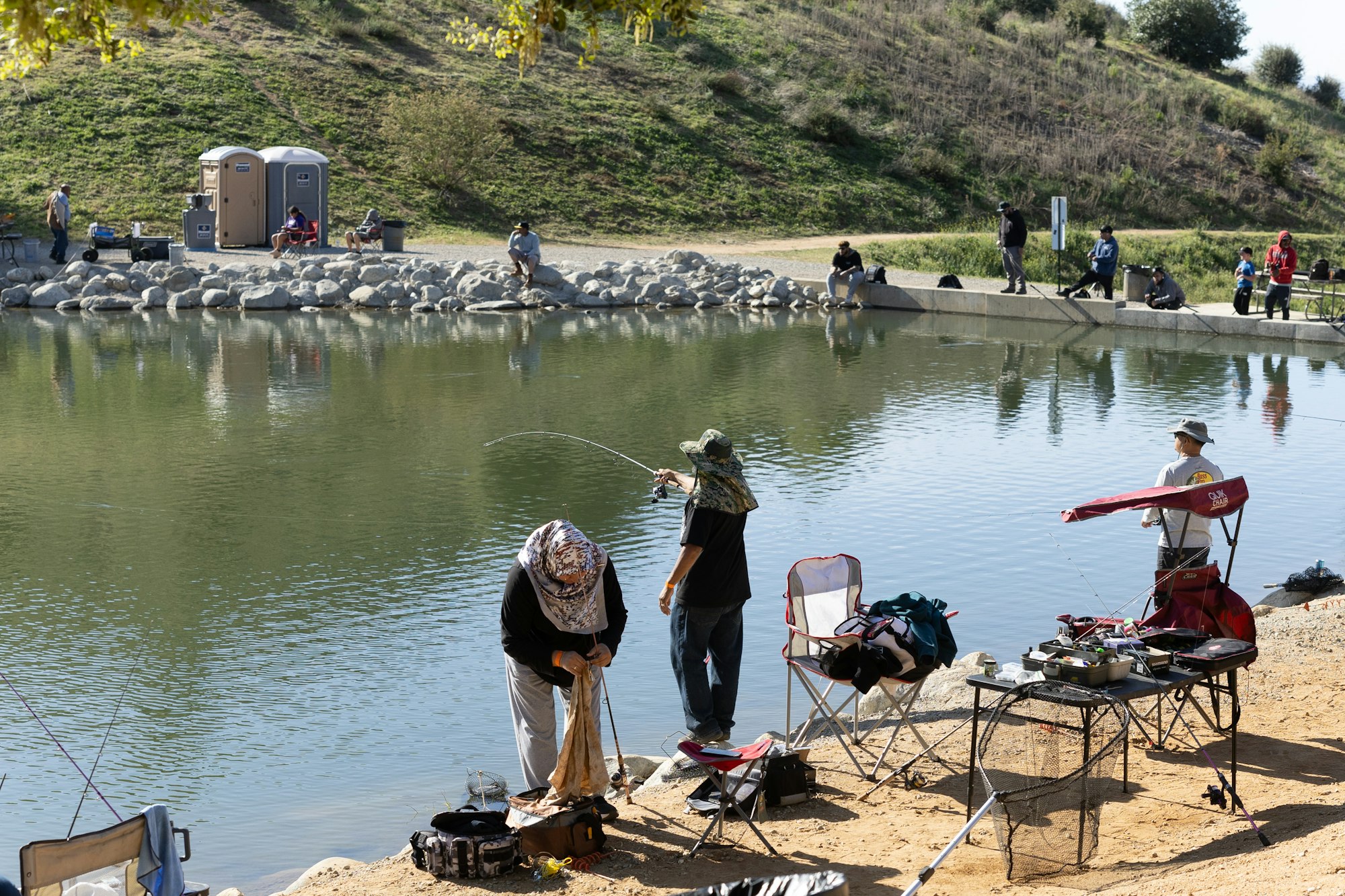 People fishing by a pond, with chairs and equipment set up, in a natural setting.