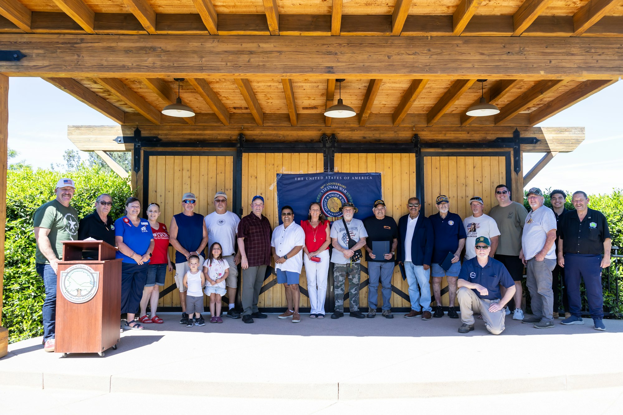 A group photo of a diverse gathering, possibly a veterans' event, posing in front of a wooden structure with a U.S. flag backdrop.