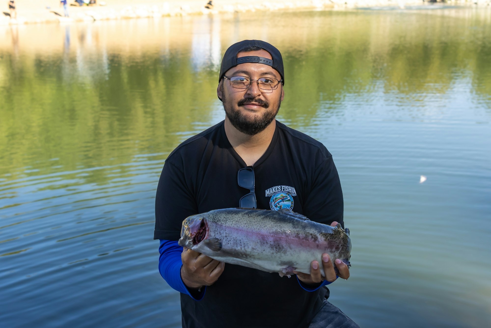 Man holding a large fish in front of a lake, wearing glasses and a cap.