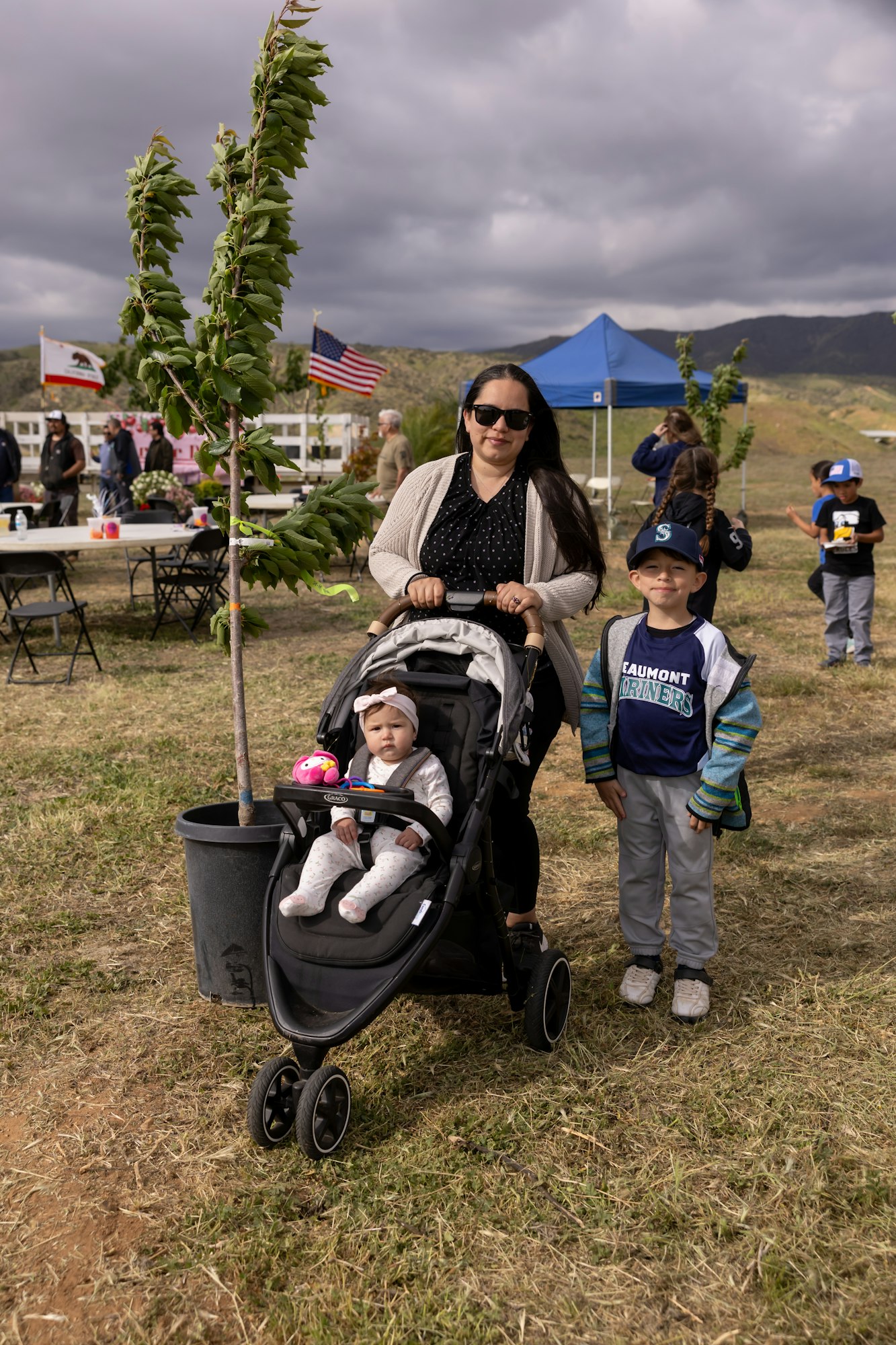 A woman with a stroller and a baby, beside a young child, at an outdoor event with flags, tents, and people in the background.