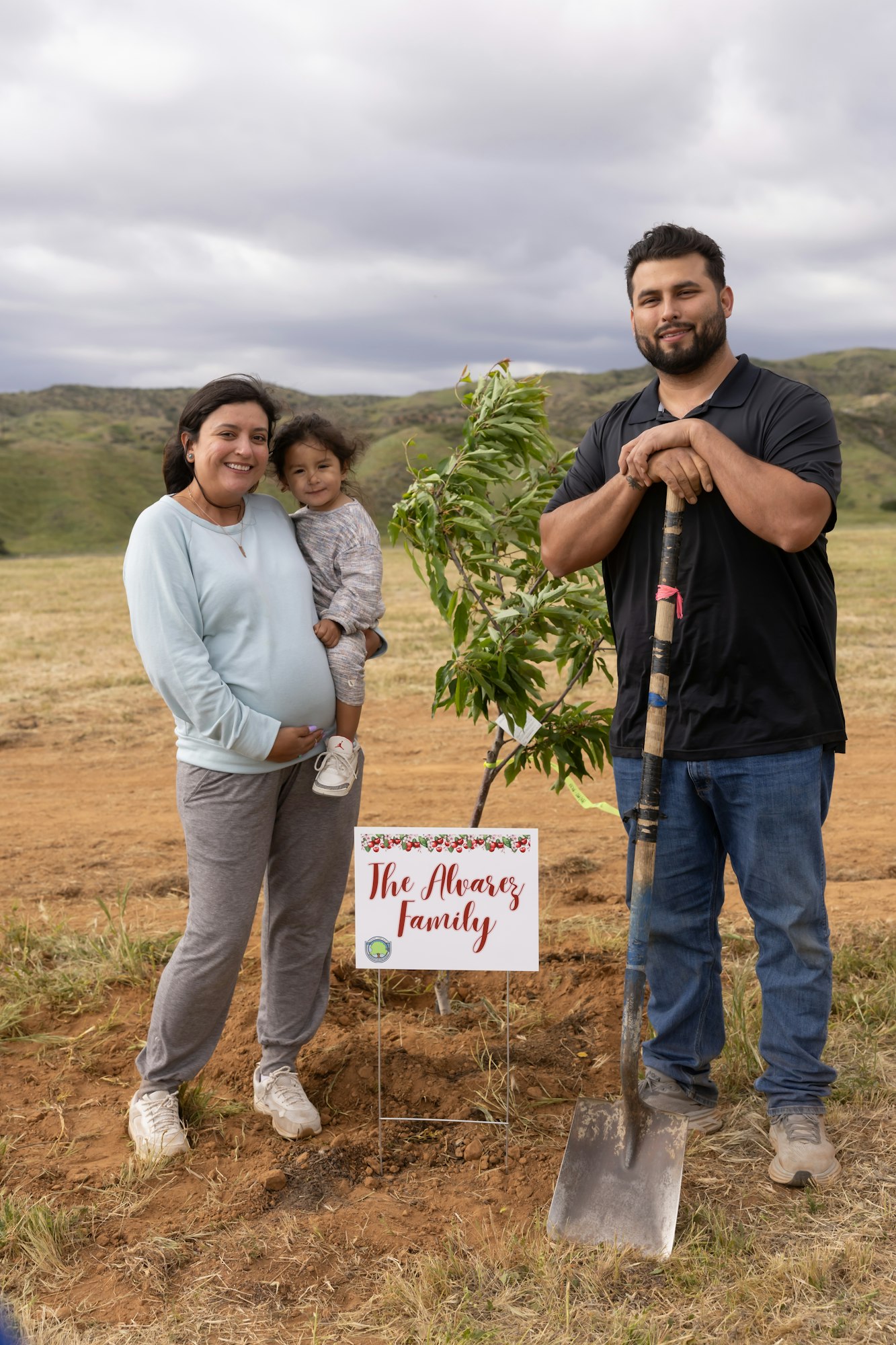 A family with a child poses beside a newly planted tree and a sign that reads "The Alvarez Family" in an open field.