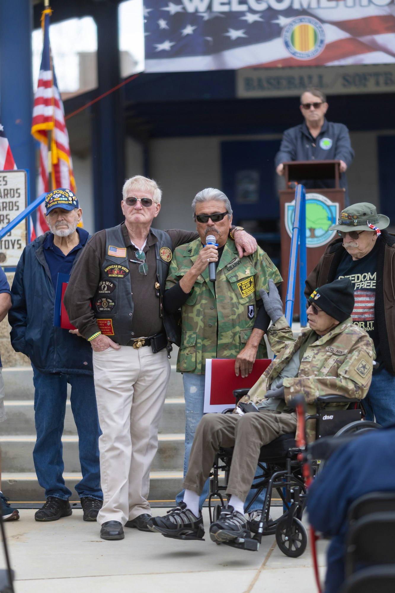 A group of veterans, some in uniforms, gather at an event. One speaks into a microphone. An American flag and "Welcome" banner are visible.