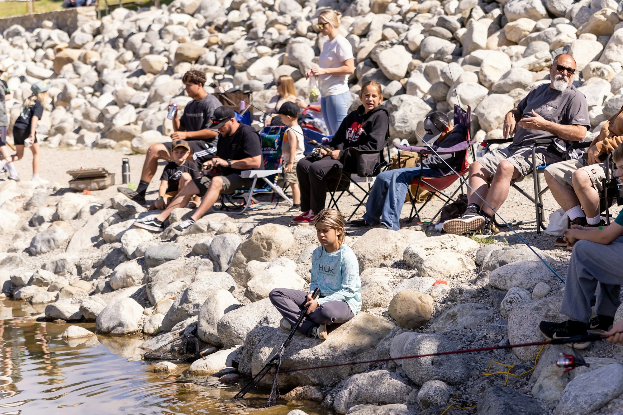 People sitting on rocky banks, fishing by the water, with various gear and chairs.