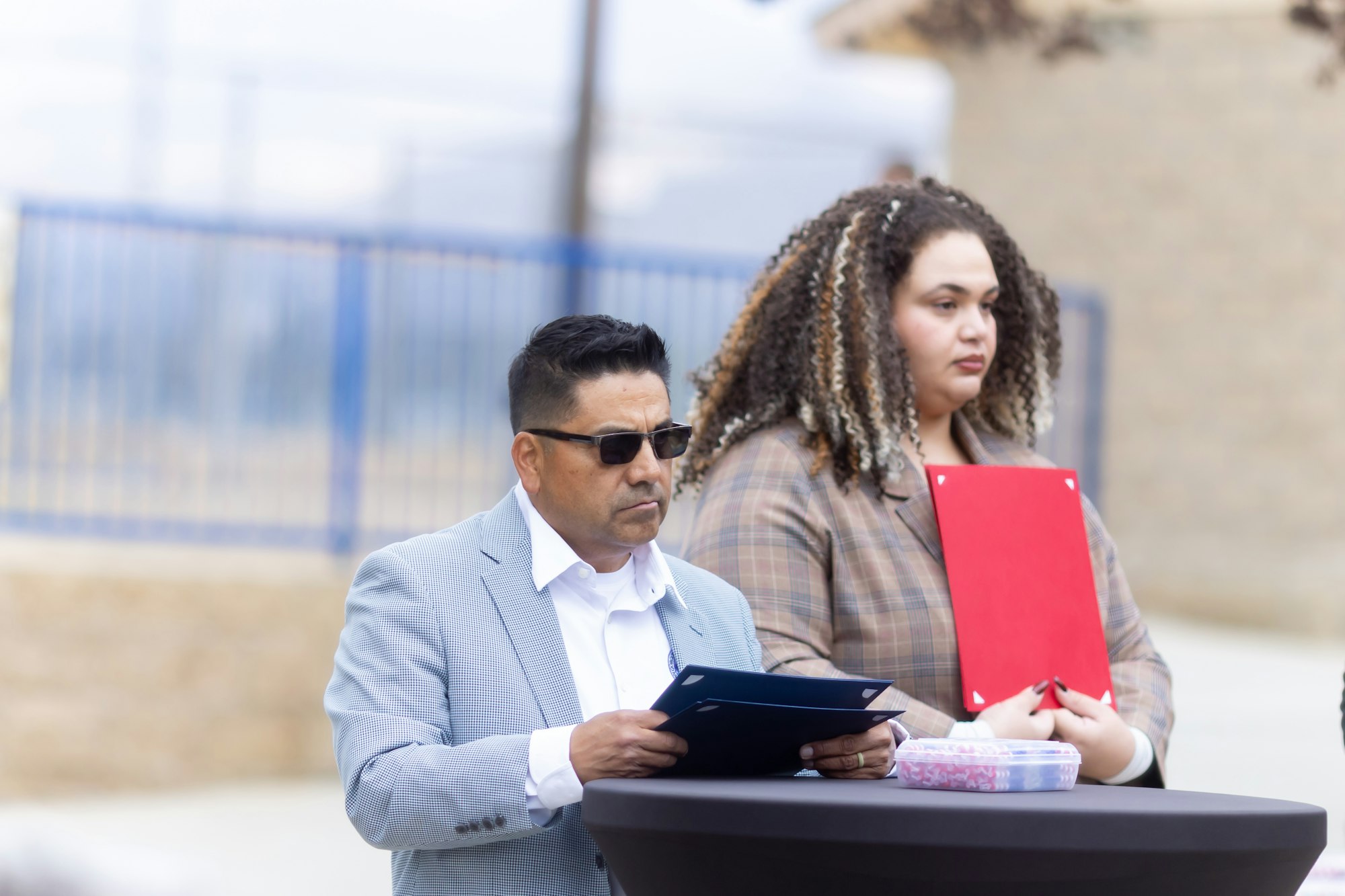 Two people holding clipboards stand by a table outdoors.