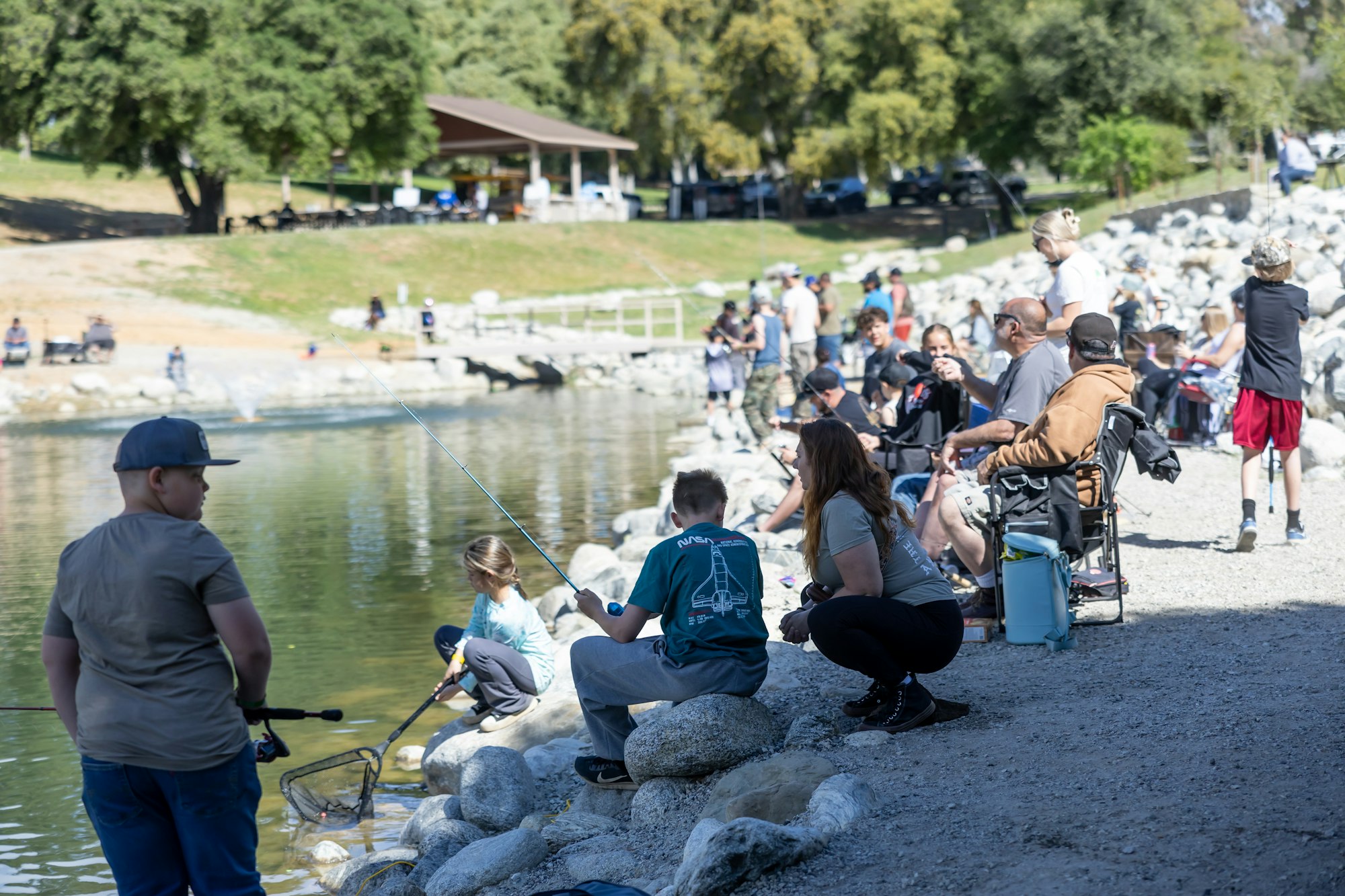 People fishing by a lakeside on a sunny day, with some sitting on rocks and others on chairs.