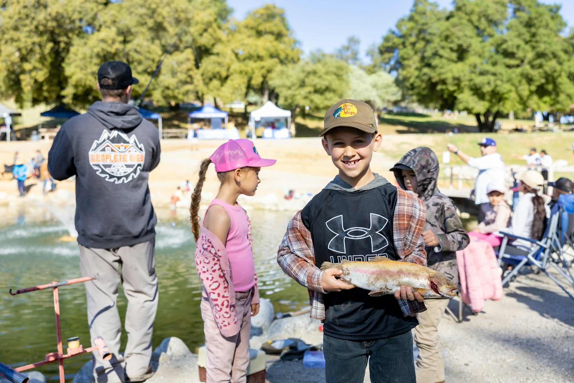A boy smiling and holding a fish, surrounded by people near a pond, outdoors on a sunny day.