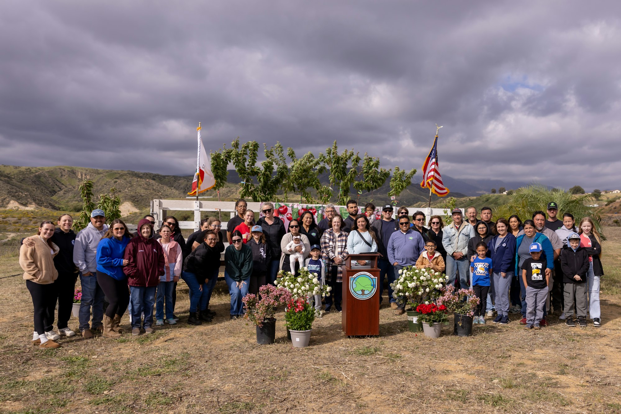 A group gathering outdoors with flags, plants, and a podium in front of a scenic hillside backdrop.