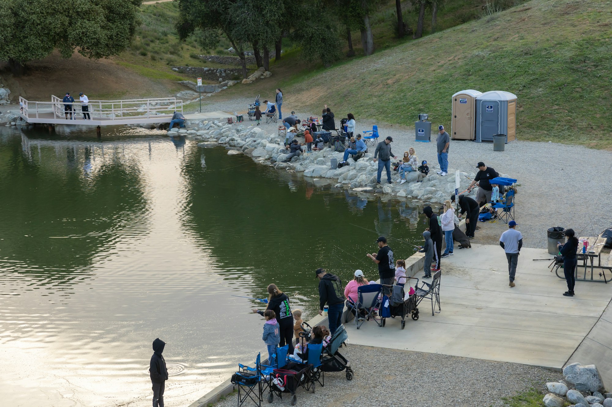 People gathered by a lakeside, fishing with gear, sitting on chairs, and socializing near portable toilets.
