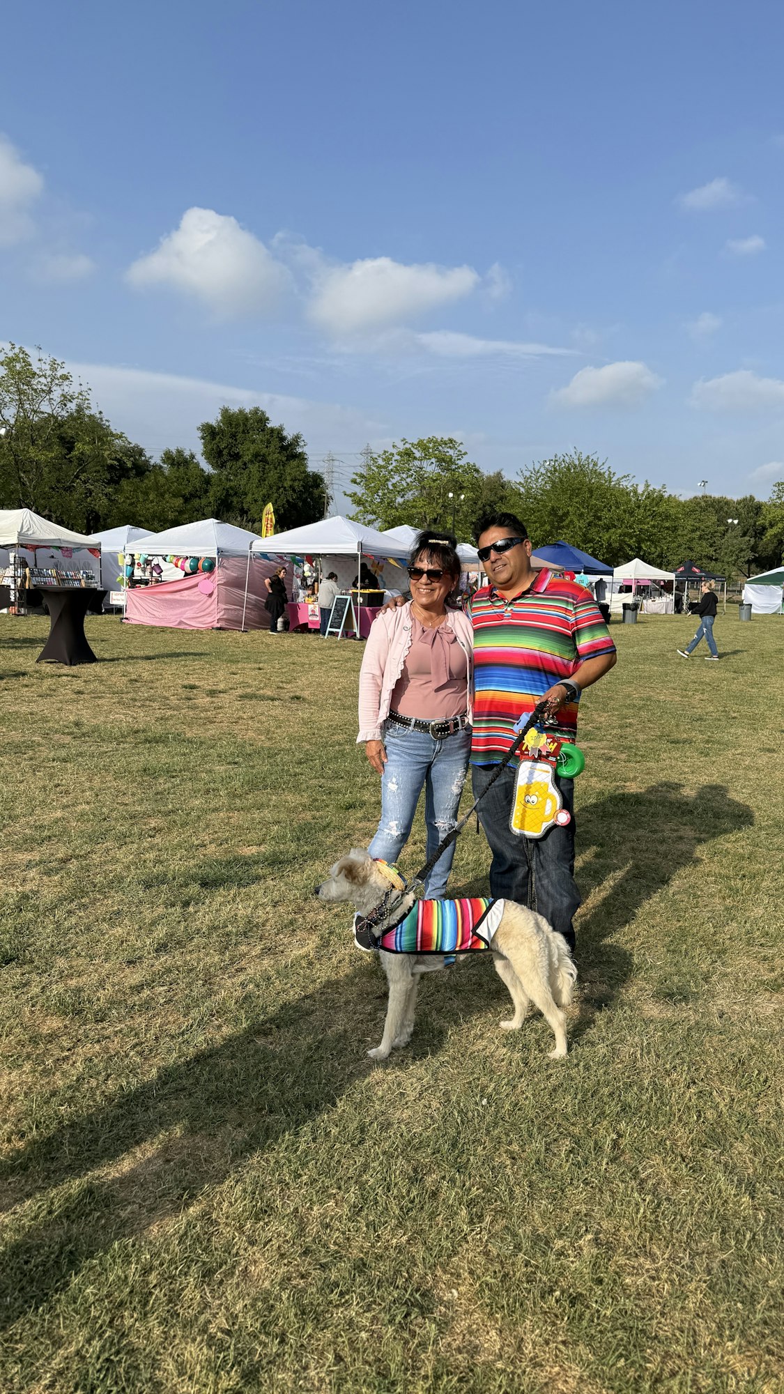 A couple poses with their dog at an outdoor festival. They wear colorful clothes, and there are tents and trees in the background.