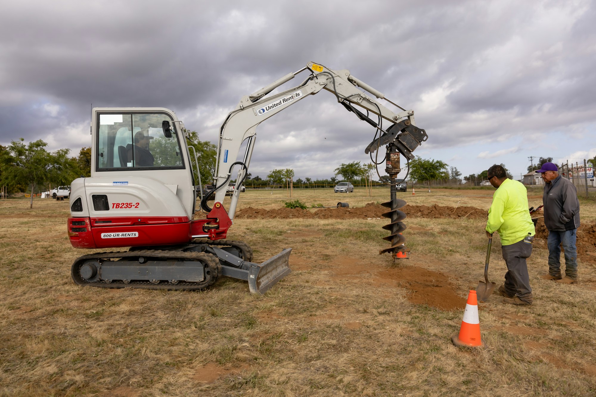 A mini excavator with an auger attachment is digging a hole while two people stand nearby on a grassy area.