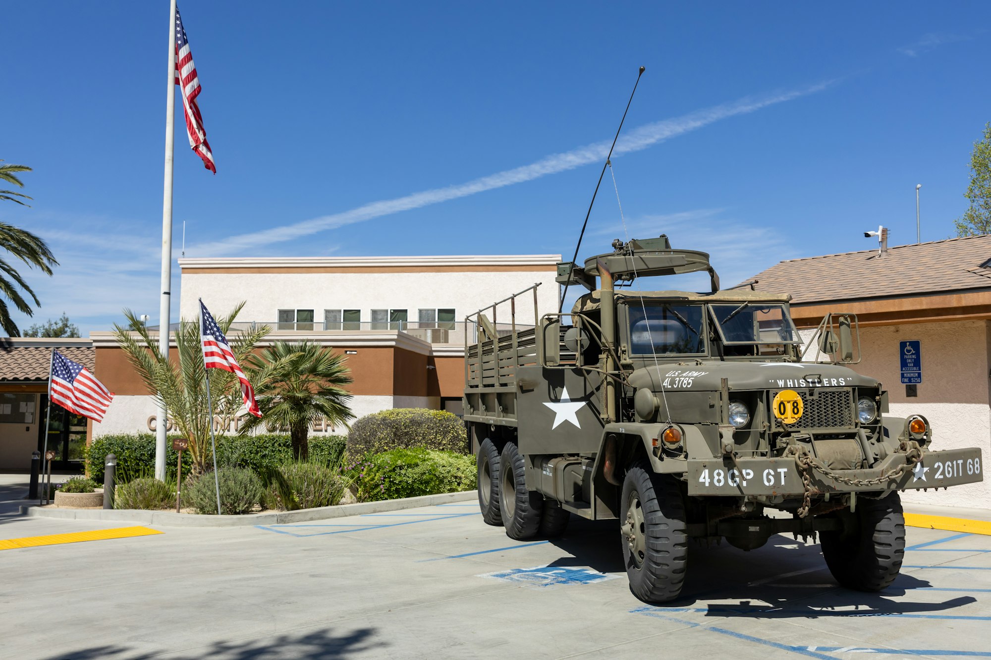 A military truck parked outside a community center, surrounded by palm trees and American flags against a clear blue sky.