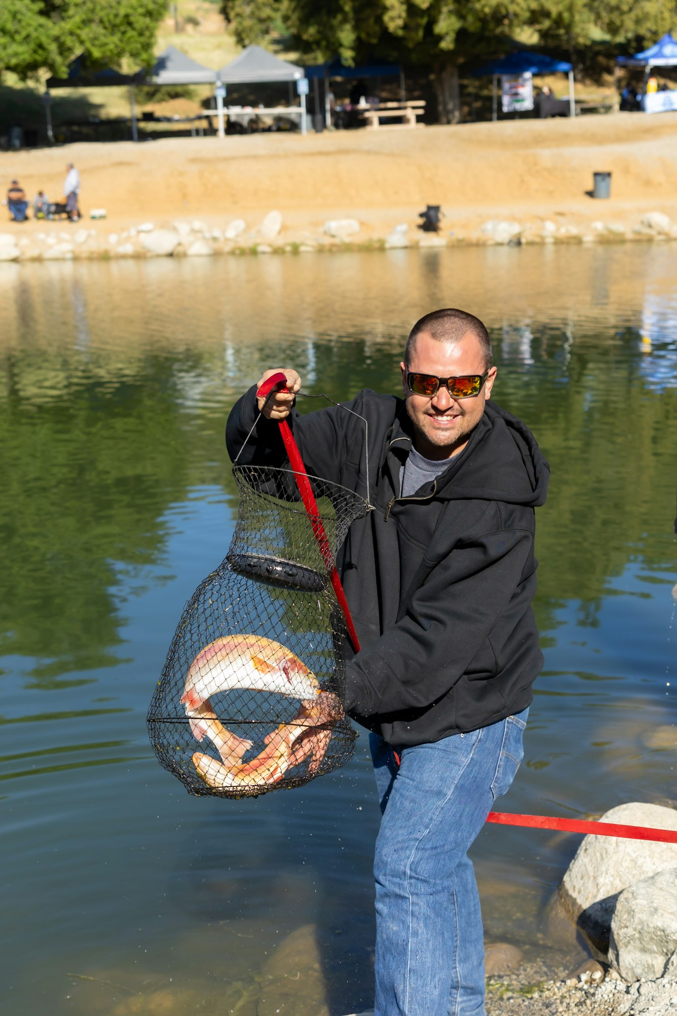 A person smiling, holding a large fish in a net by a lake, wearing sunglasses and a hoodie.