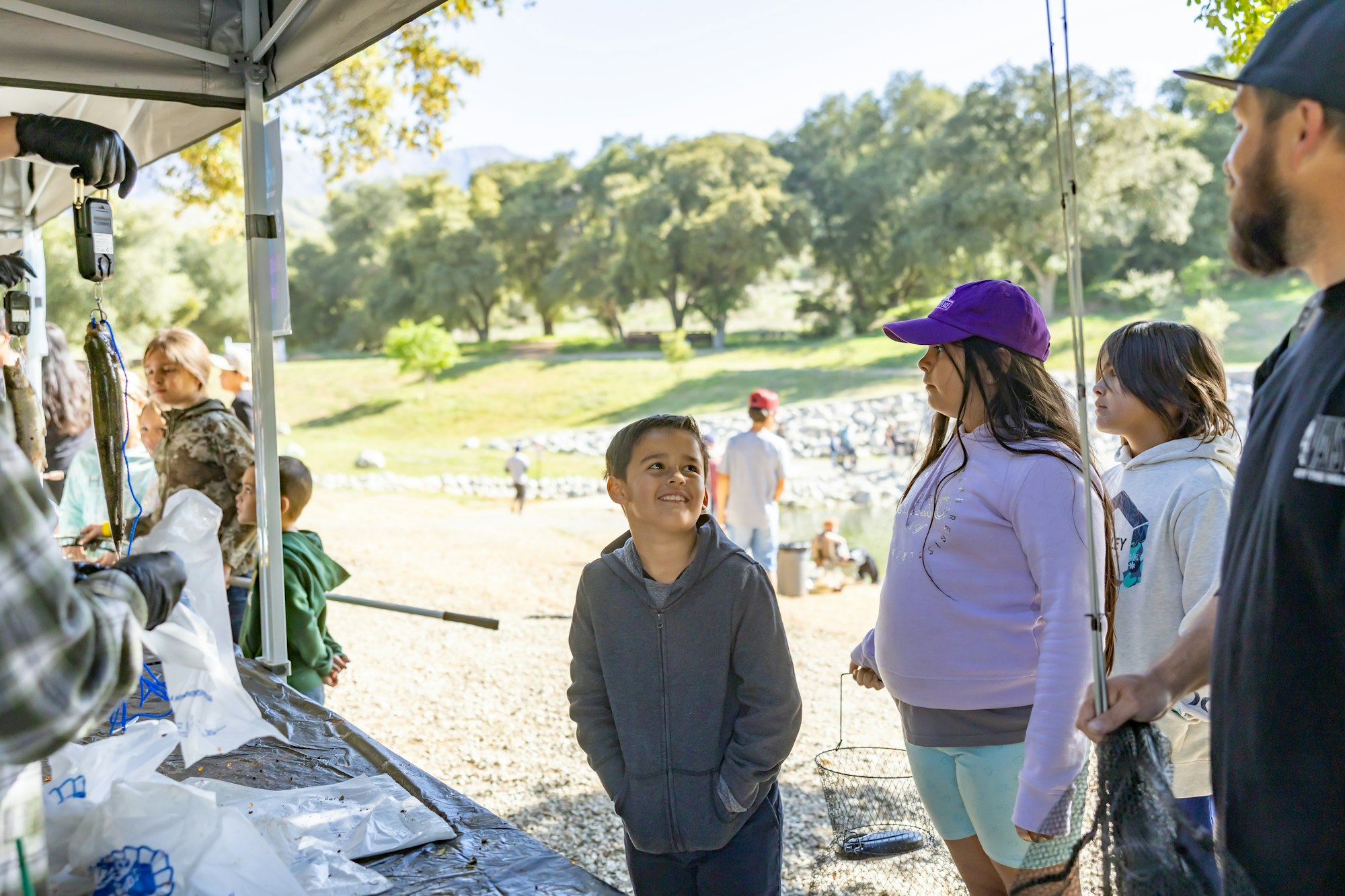 People standing at a tent, one holds a fish. A child smiles, others hold fishing gear. Outdoor setting with trees in the background.