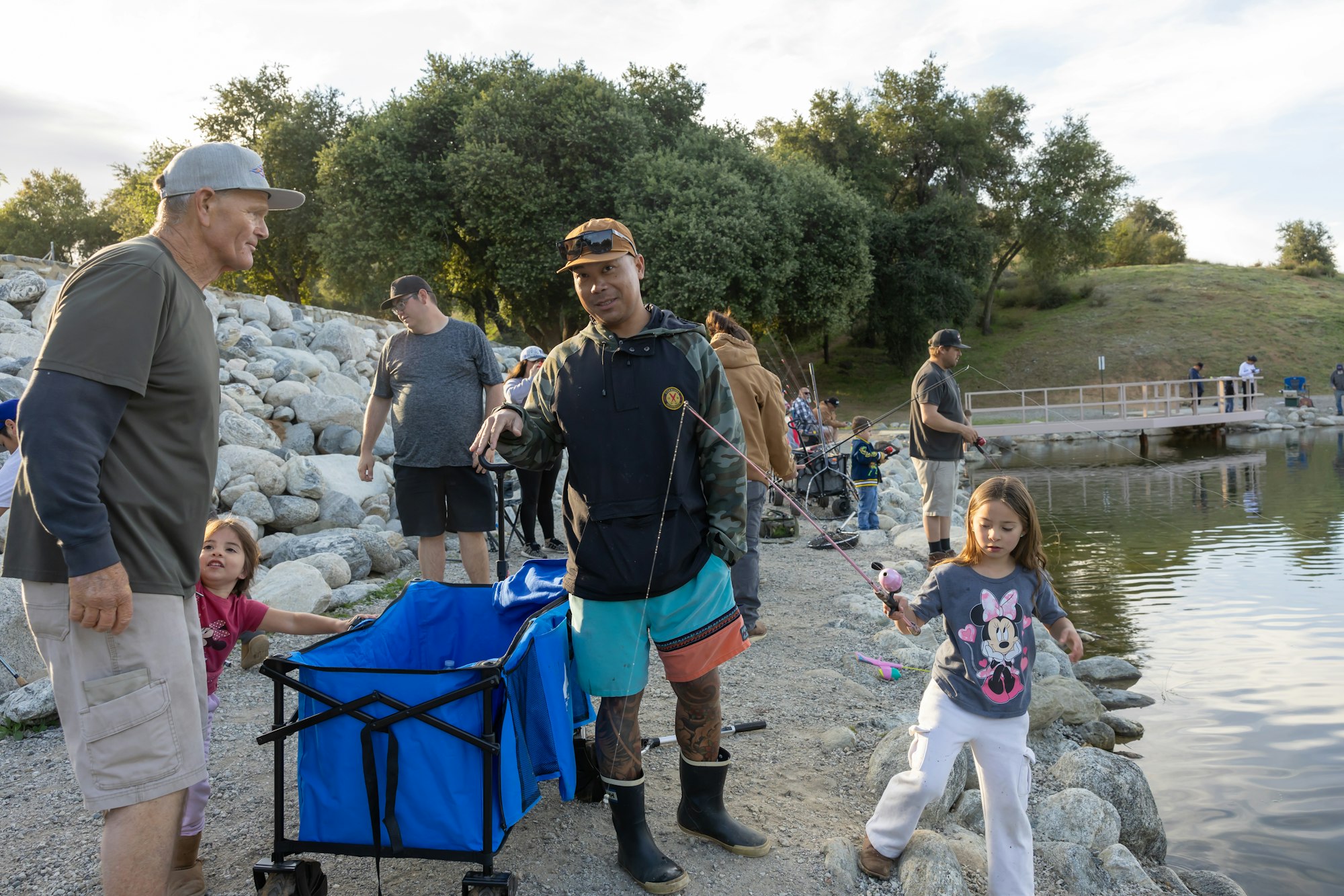 People fishing by a lake, including adults and children, with one person wearing boots holding a fishing rod near a blue cart.