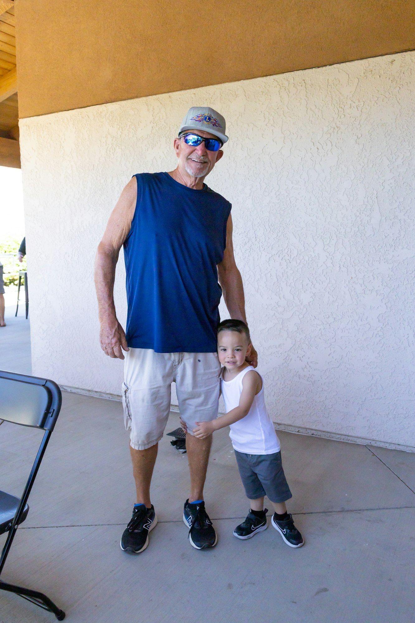 A smiling older man in a navy tank top and sunglasses stands with a young boy in a white tank top, both looking happy together.