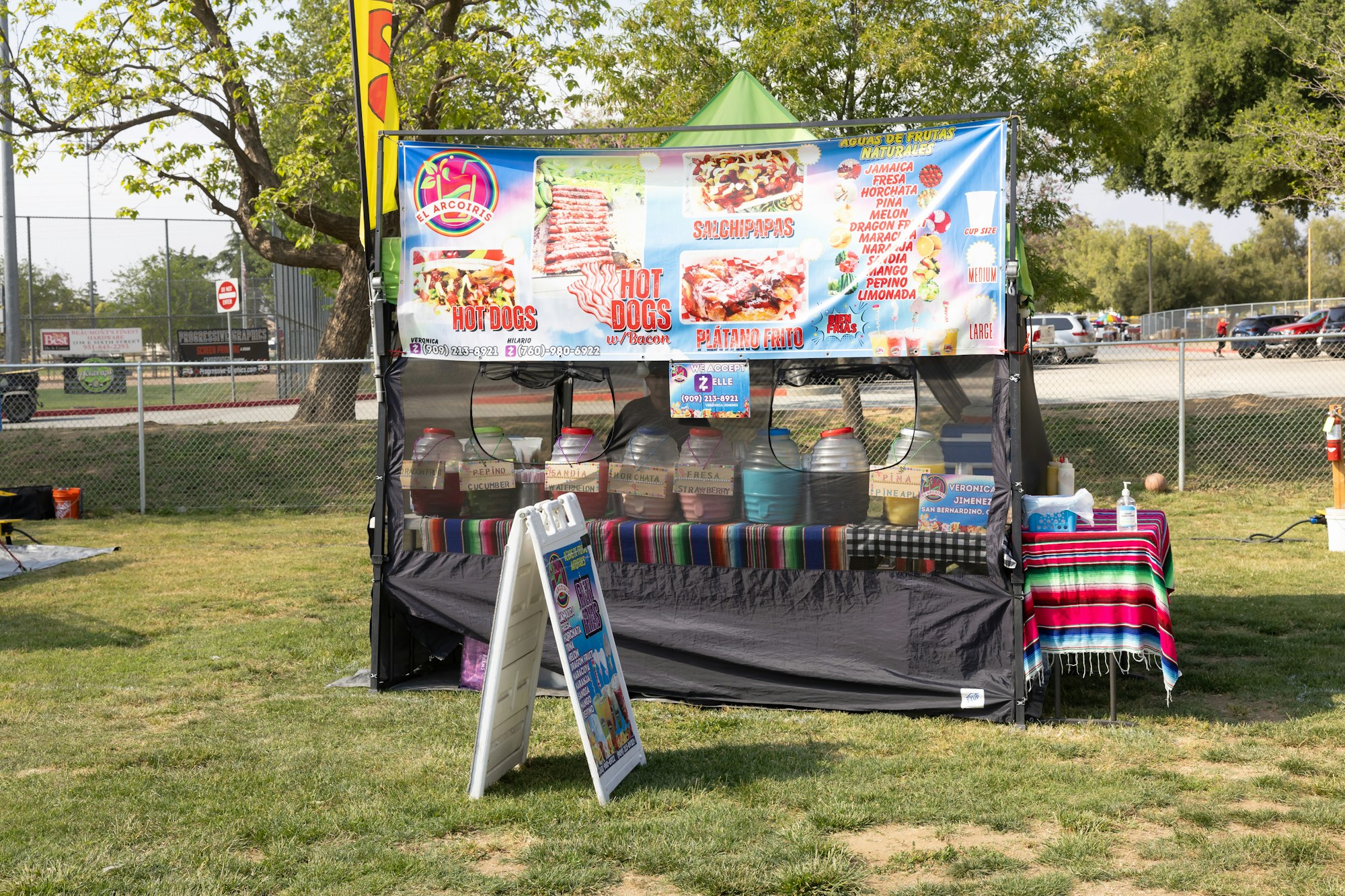 Outdoor food stand selling hot dogs, aguas frescas, and snacks, with colorful signage and jugs of drinks on display.
