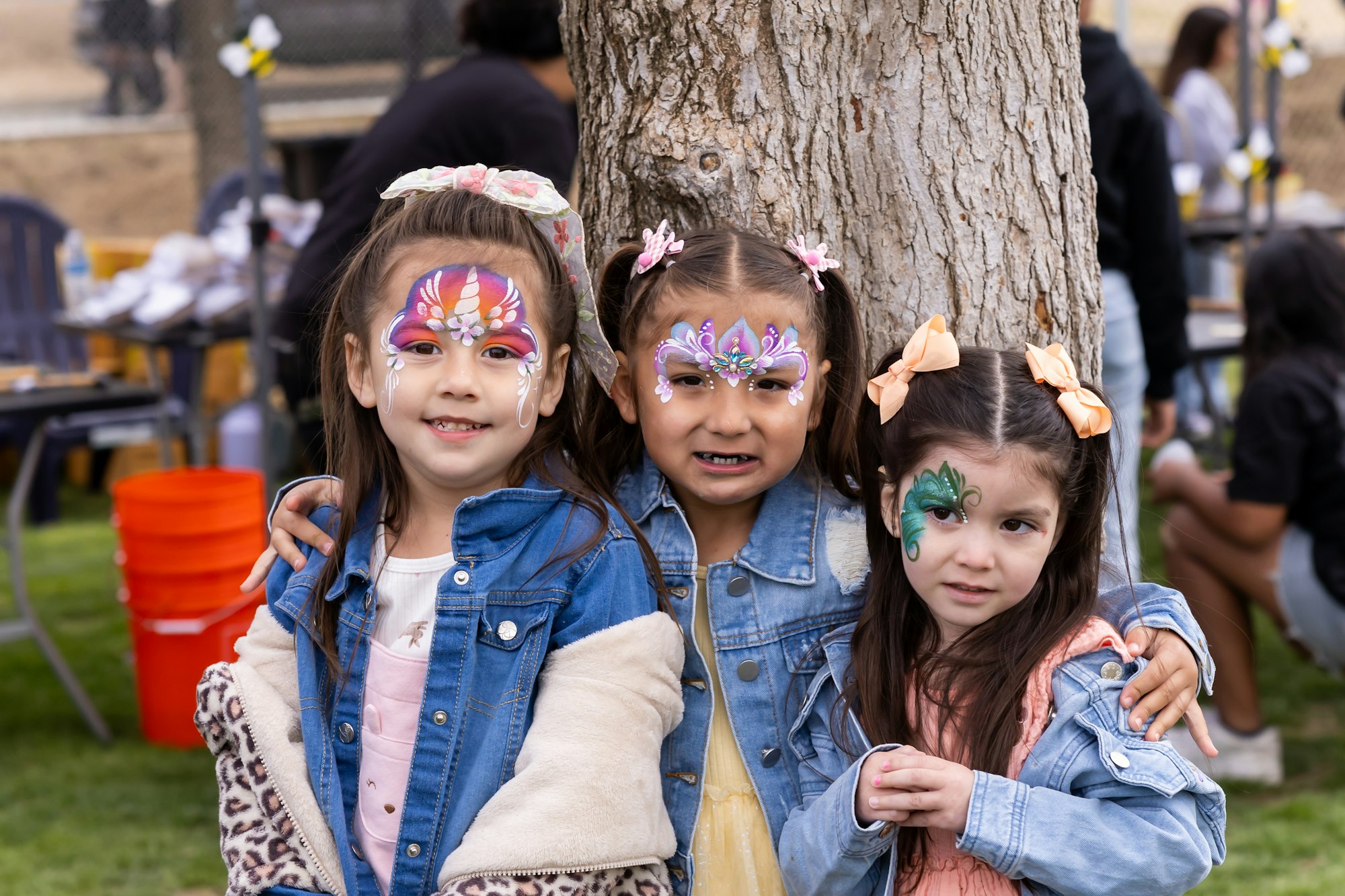 Three children with face paint and denim jackets standing together outside by a tree.