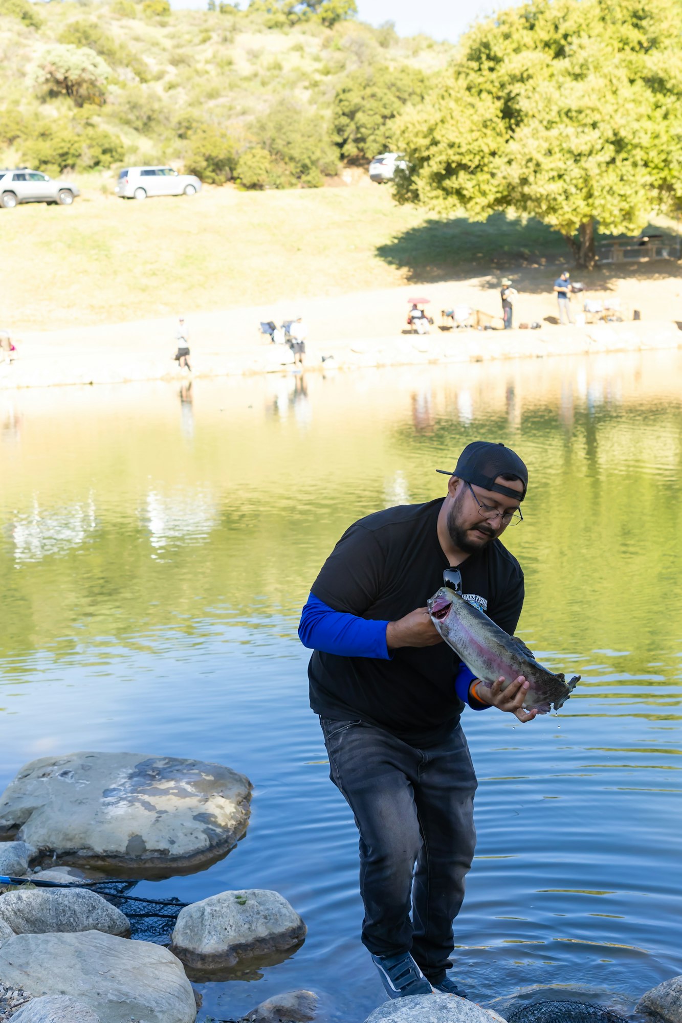 Man by a lake holding a large fish, with people and cars in the background.
