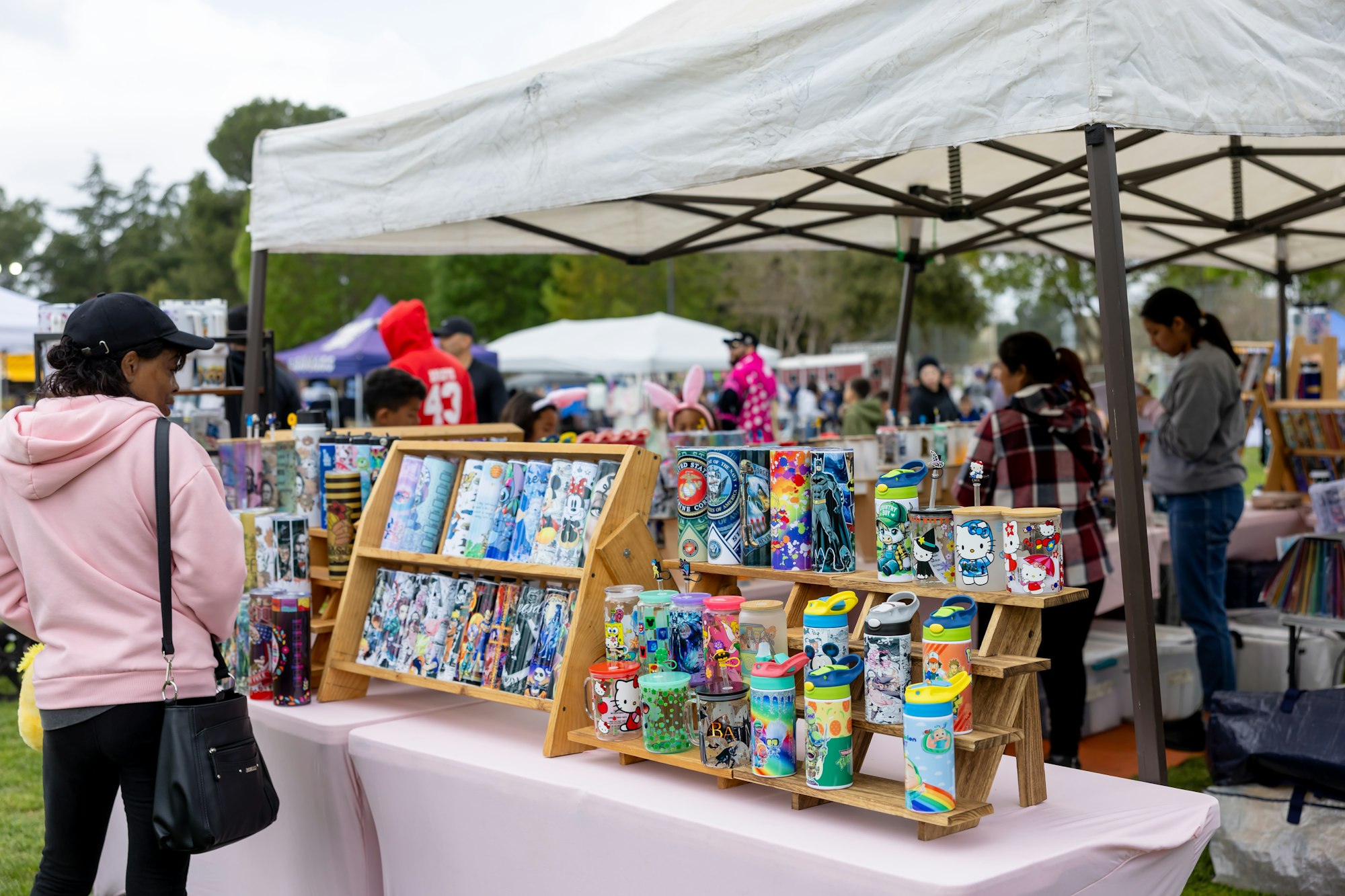 Outdoor market stall with colorful tumblers and cups, people browsing and shopping under a canopy.