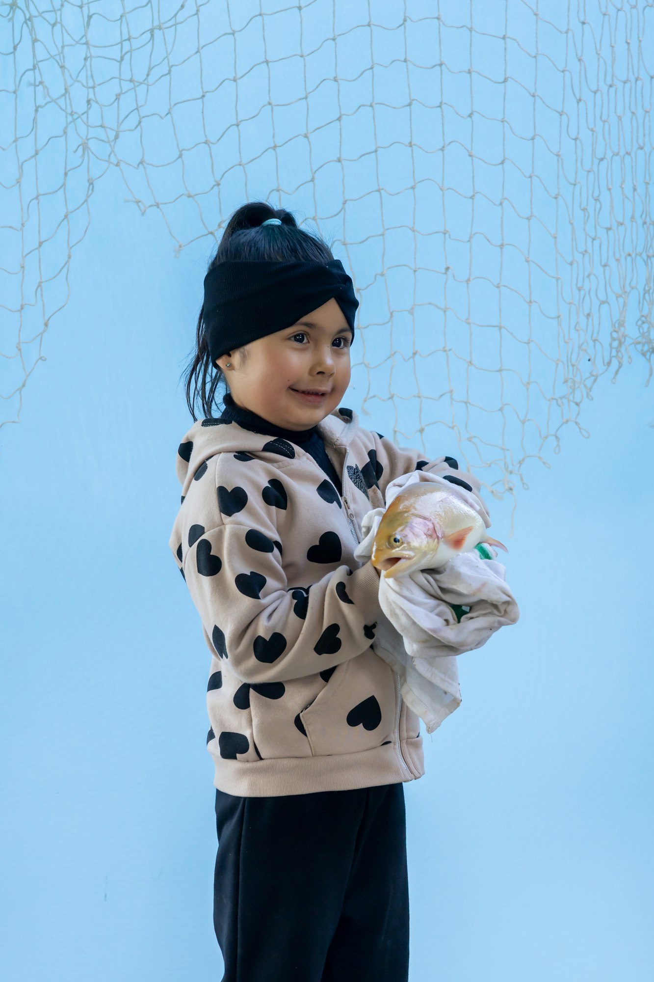 A child in a heart-patterned hoodie holds a fish wrapped in a cloth against a blue background with a net.