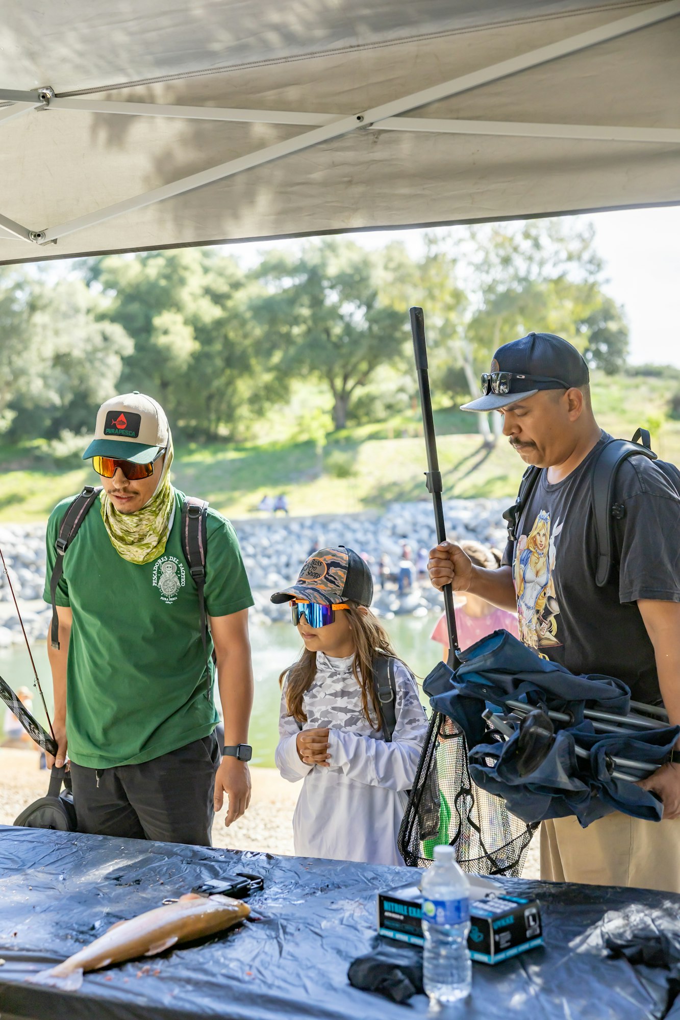 Three people with fishing gear stand by a table with a fish and water bottle outdoors.