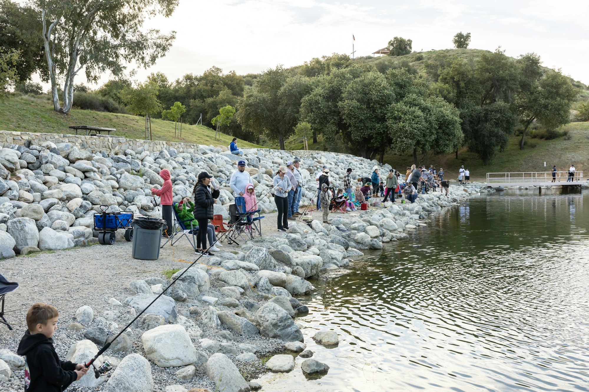 People fishing along a rocky lakeside with trees and grassy hills in the background.