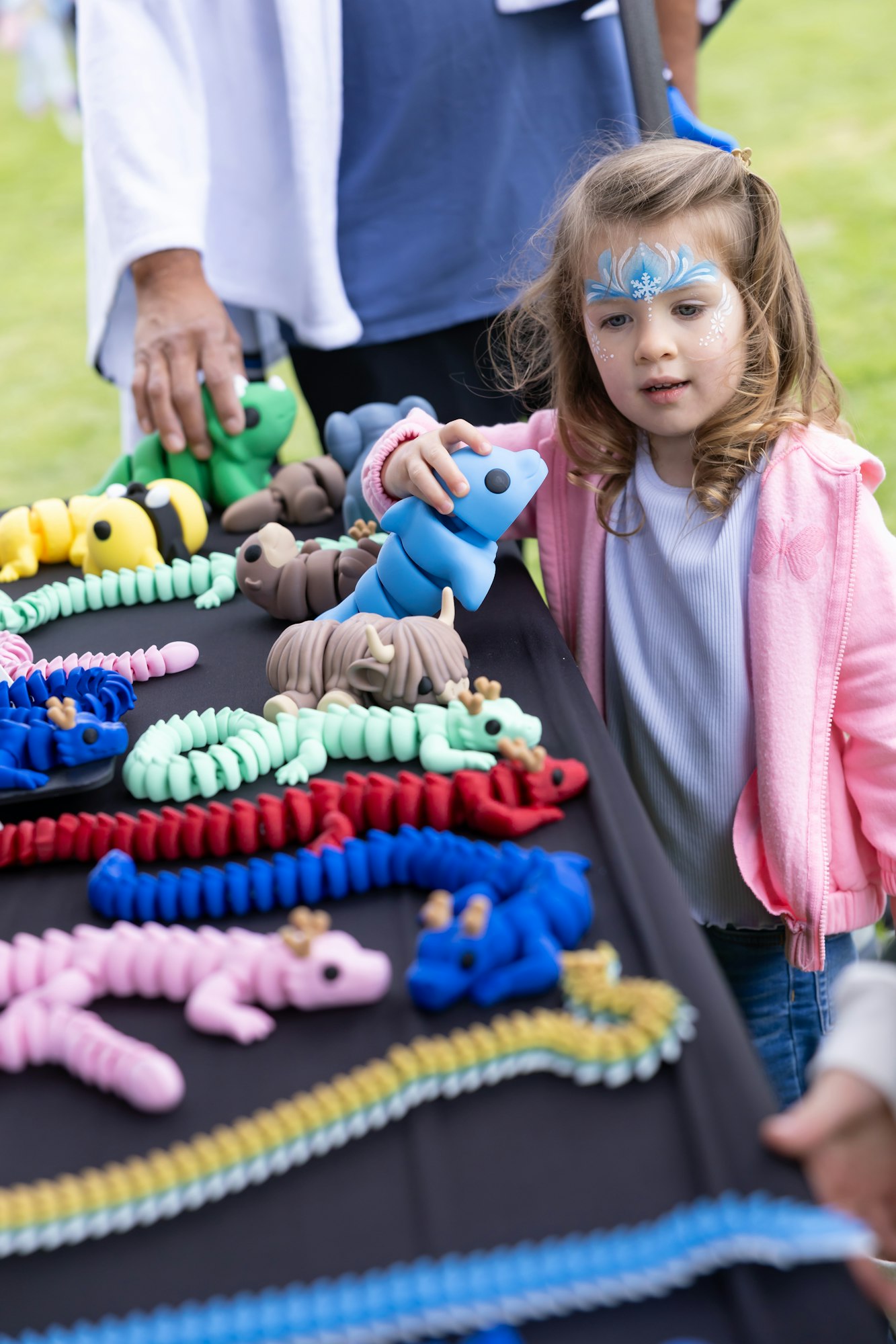 A child with face paint examines colorful toy animals at a table display outdoors.