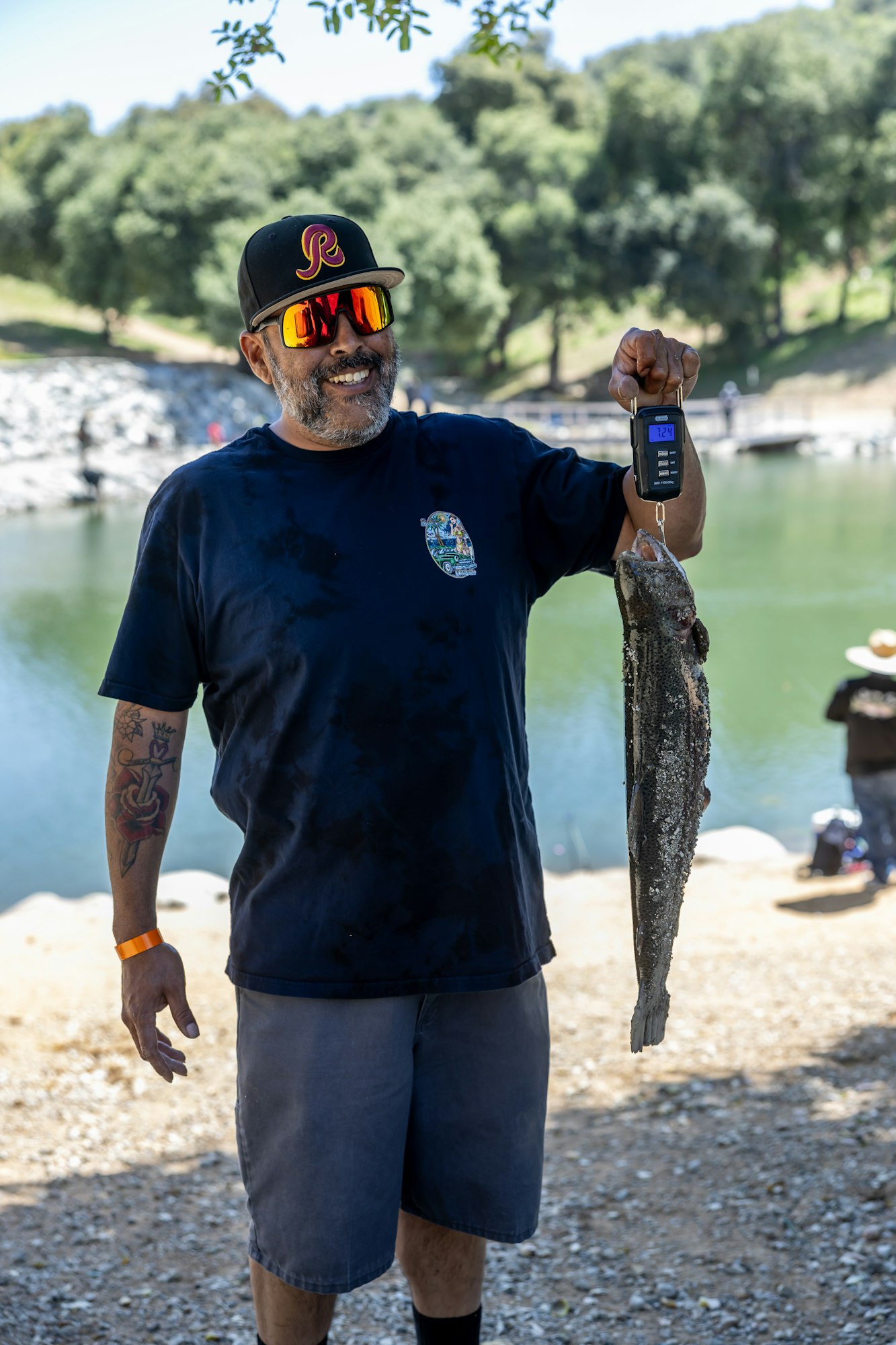 A person smiling, holding a fish on a digital scale outdoors near a lake.