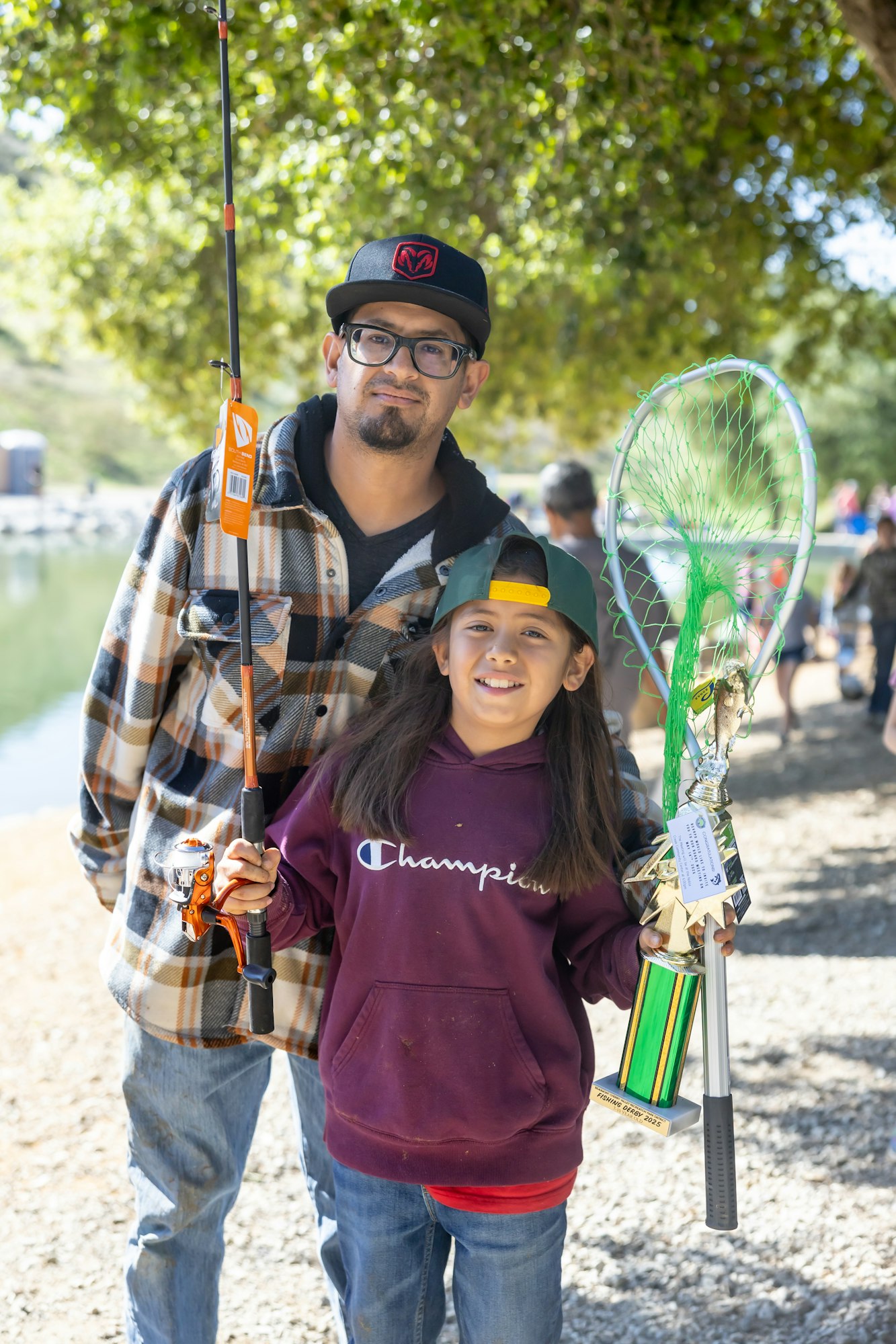 Person and a child with fishing gear and a trophy, outdoors by a lake.