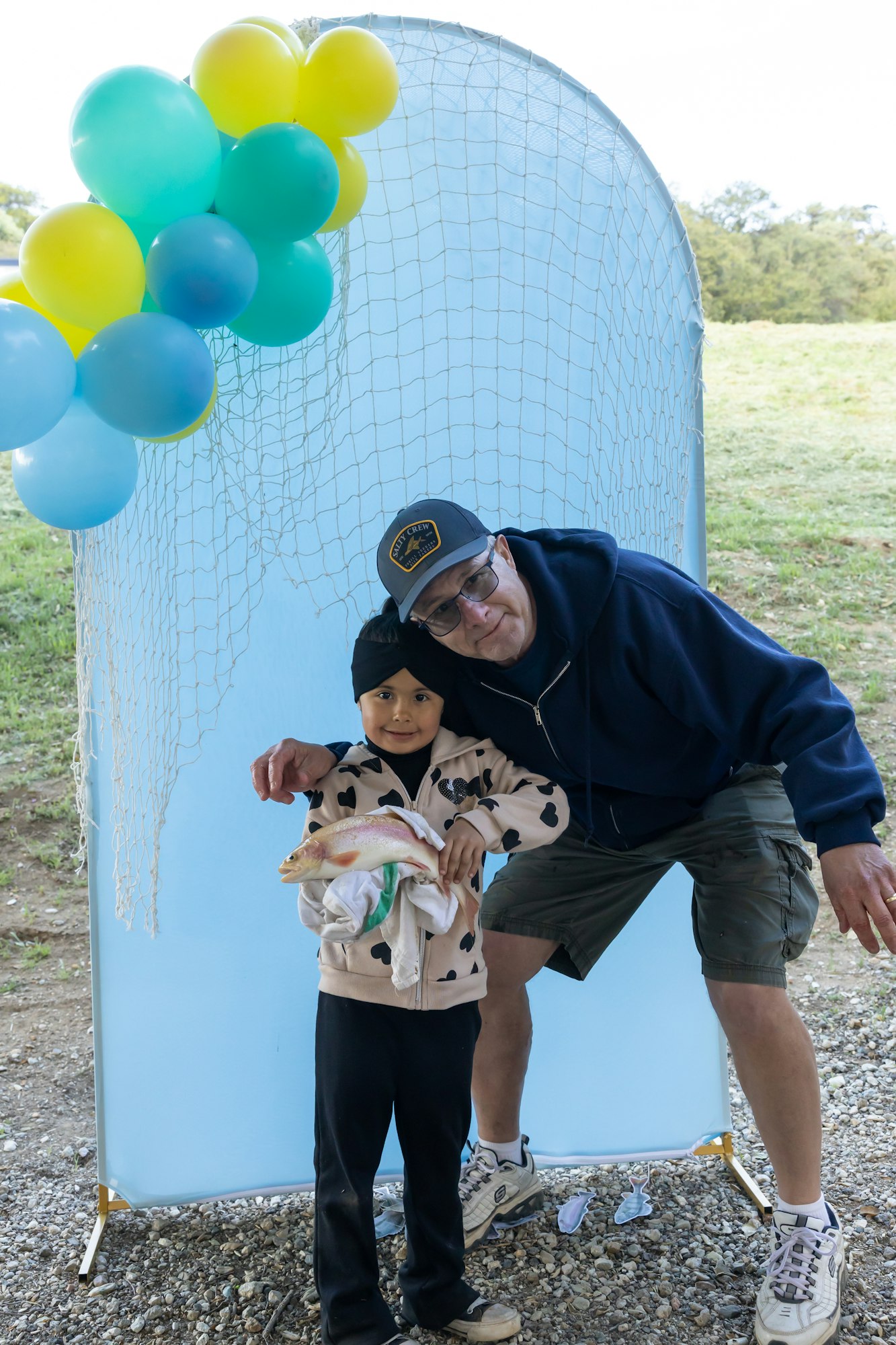 Child and adult posing with a fish, balloons, and a blue net backdrop outdoors.