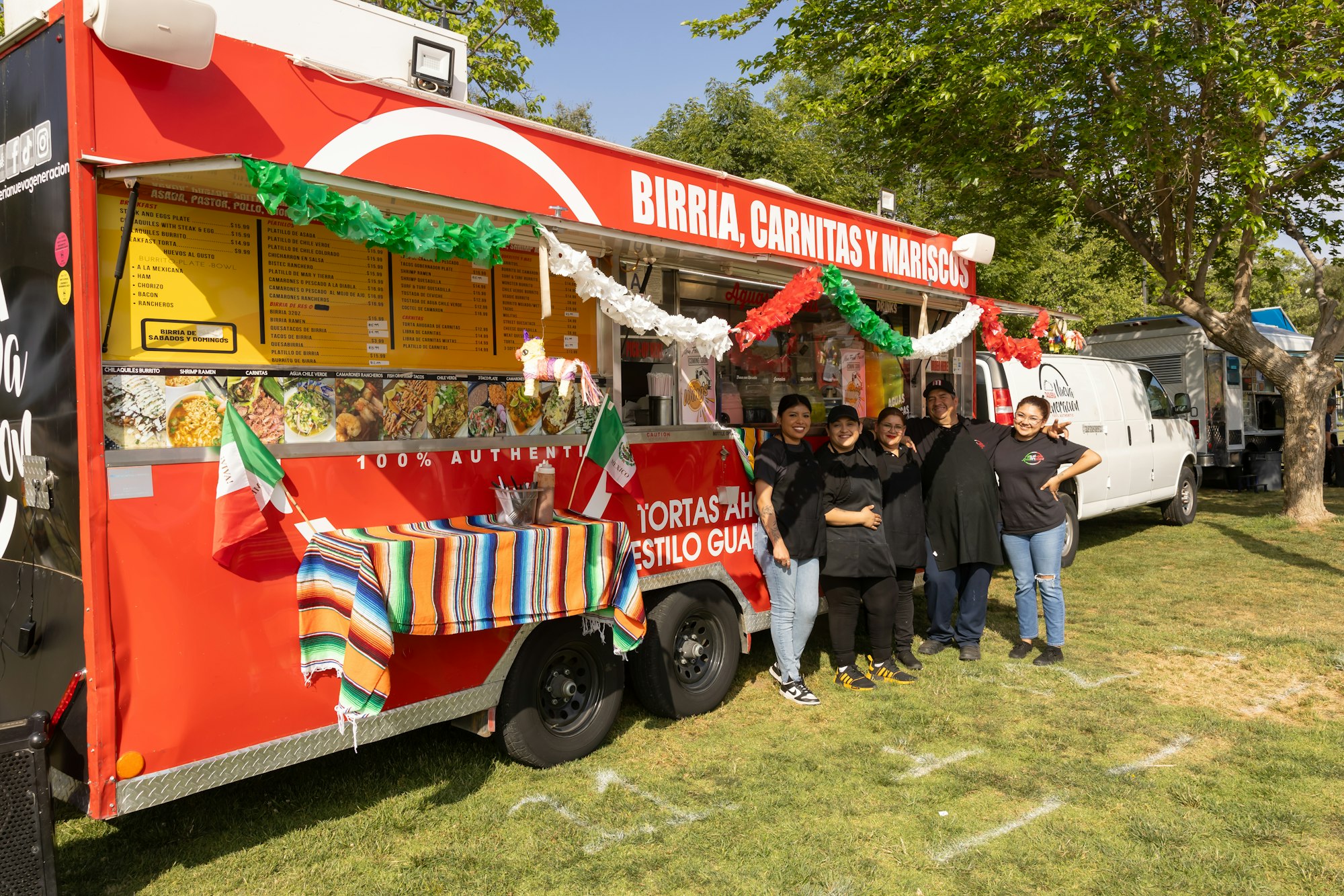 A red food truck labeled "BIRRIA, CARNITAS Y MARISCOS" with a group of people standing in front, adorned with Mexican flags and decorations.