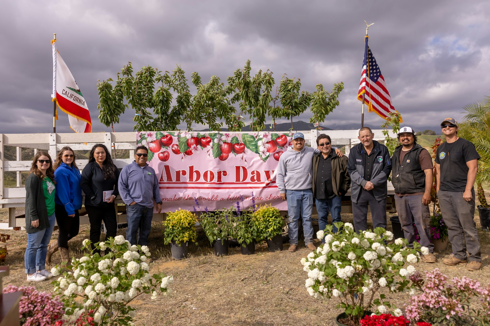 Group posing by "Arbor Day" sign with flowers, trees, and California and US flags in the background.