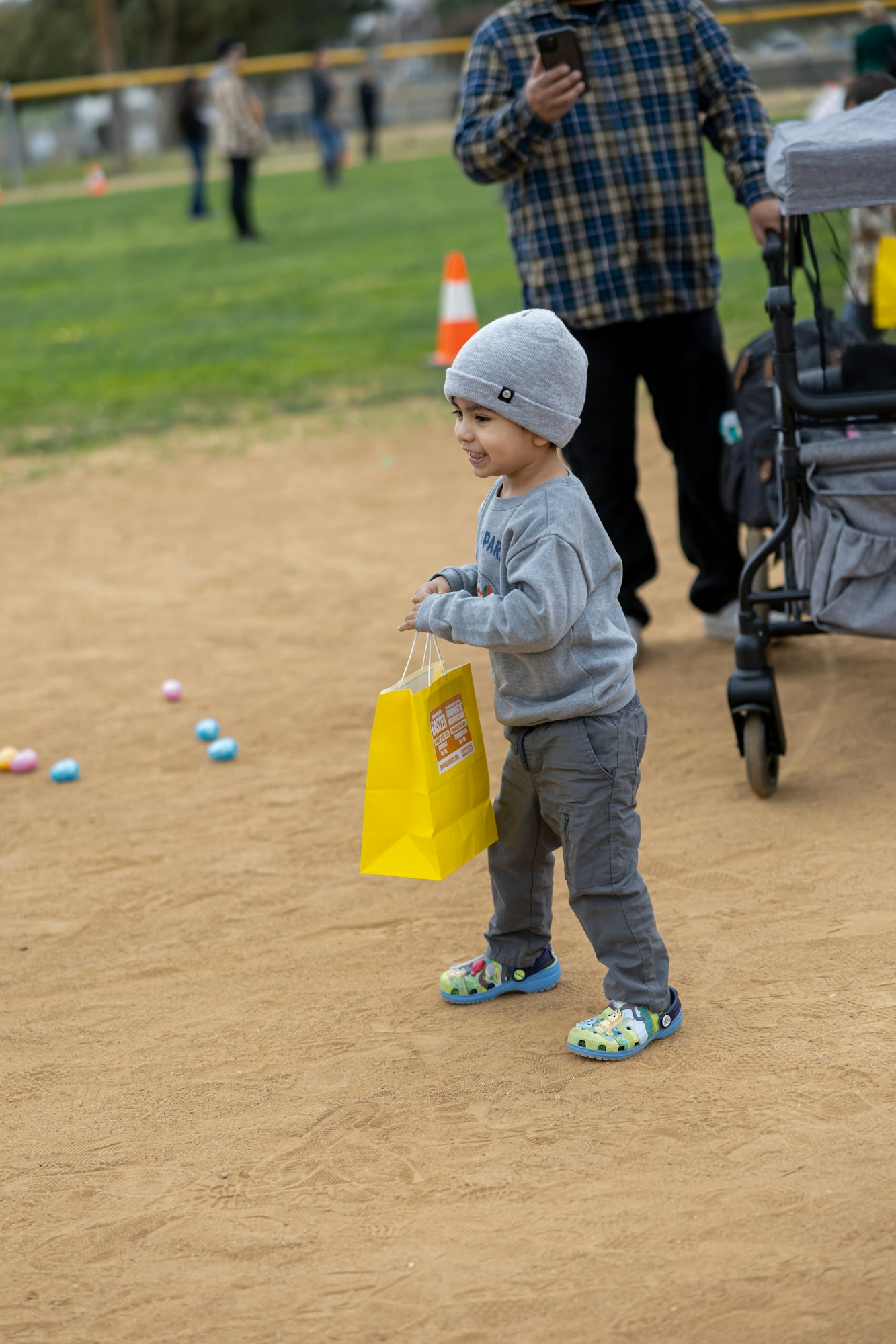 A child wearing a gray beanie and holding a yellow bag stands on a dirt path near scattered colorful eggs.