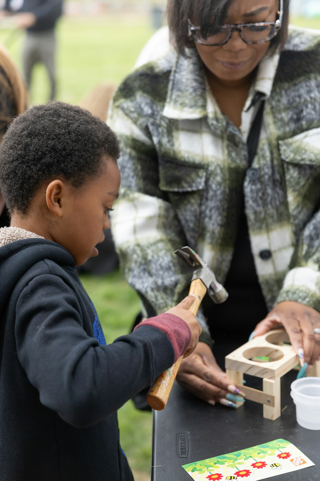 Child hammering a nail into a small wooden project, assisted by an adult.