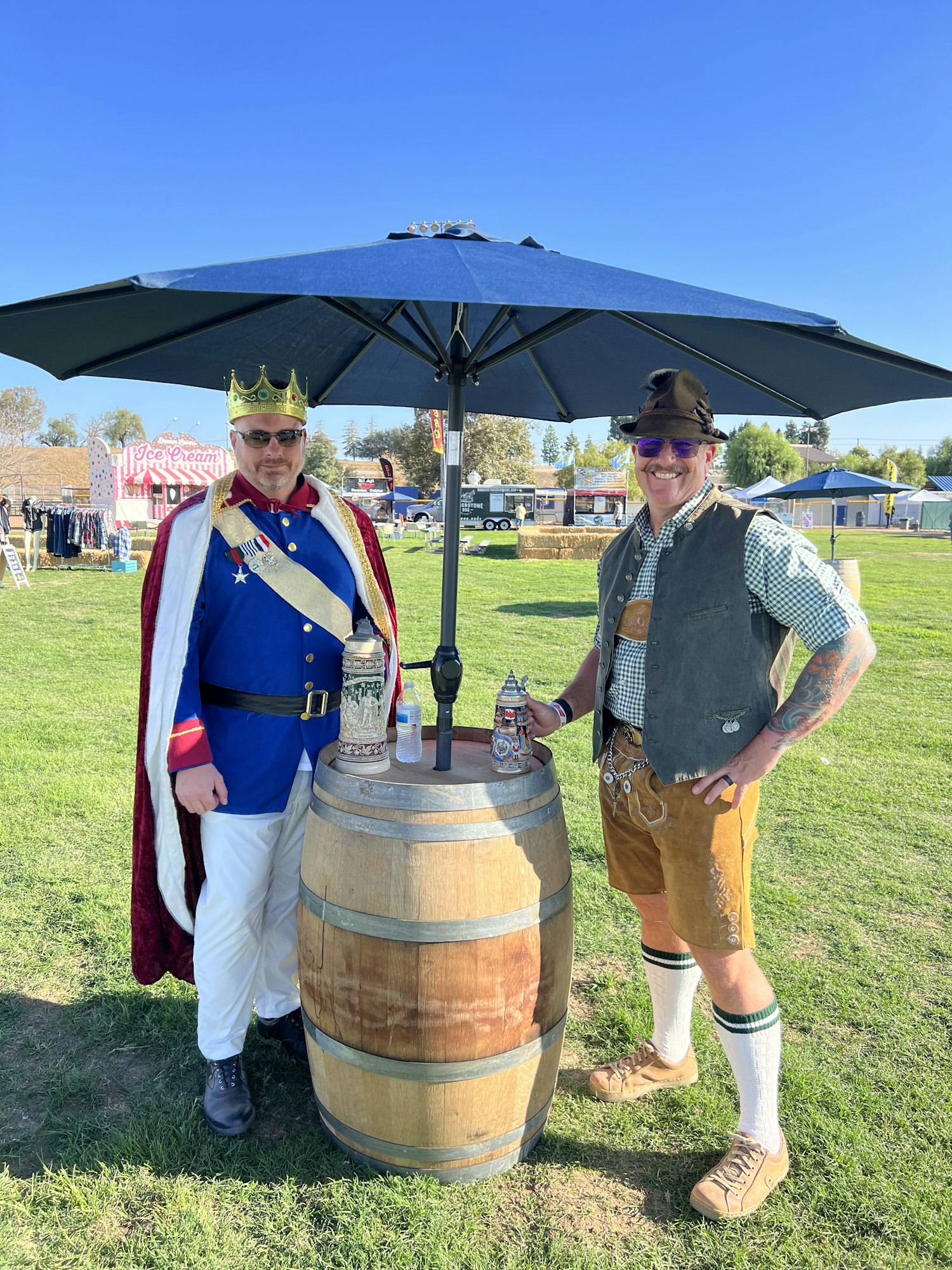 Two people in costume stand by a barrel under an umbrella on a grassy field.
