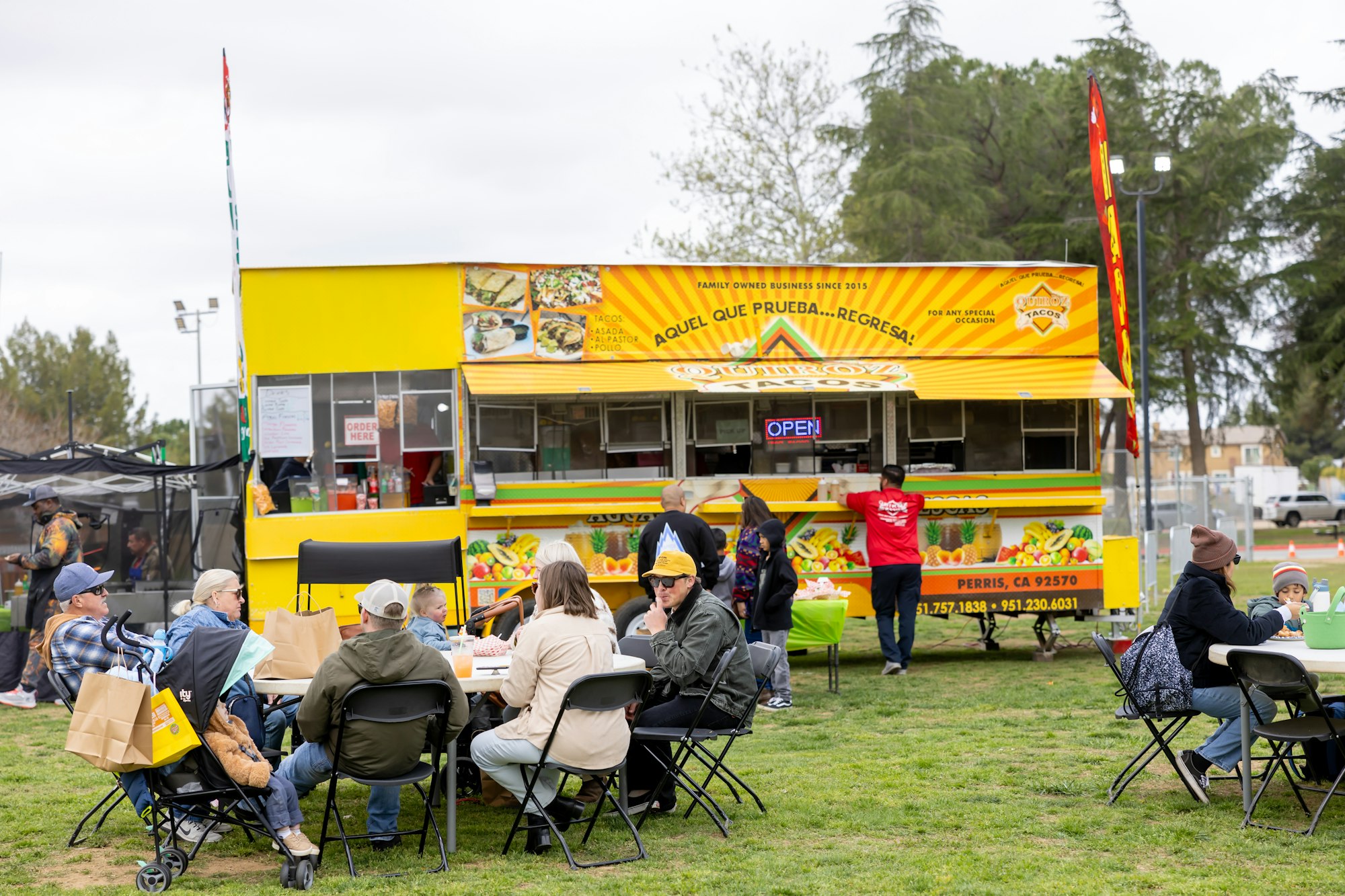 A food truck event with people sitting and eating at tables on a grass field.