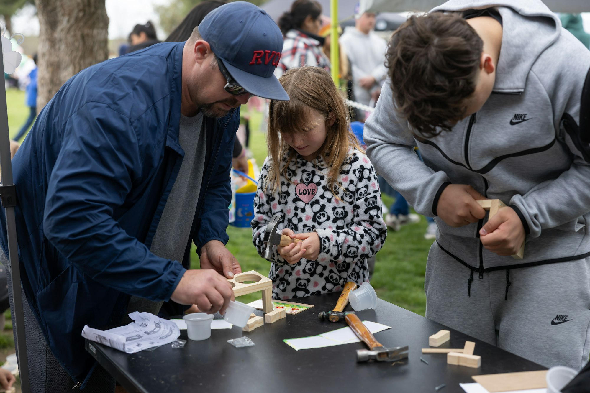 Three people at an outdoor table, assembling wooden crafts with tools and materials.