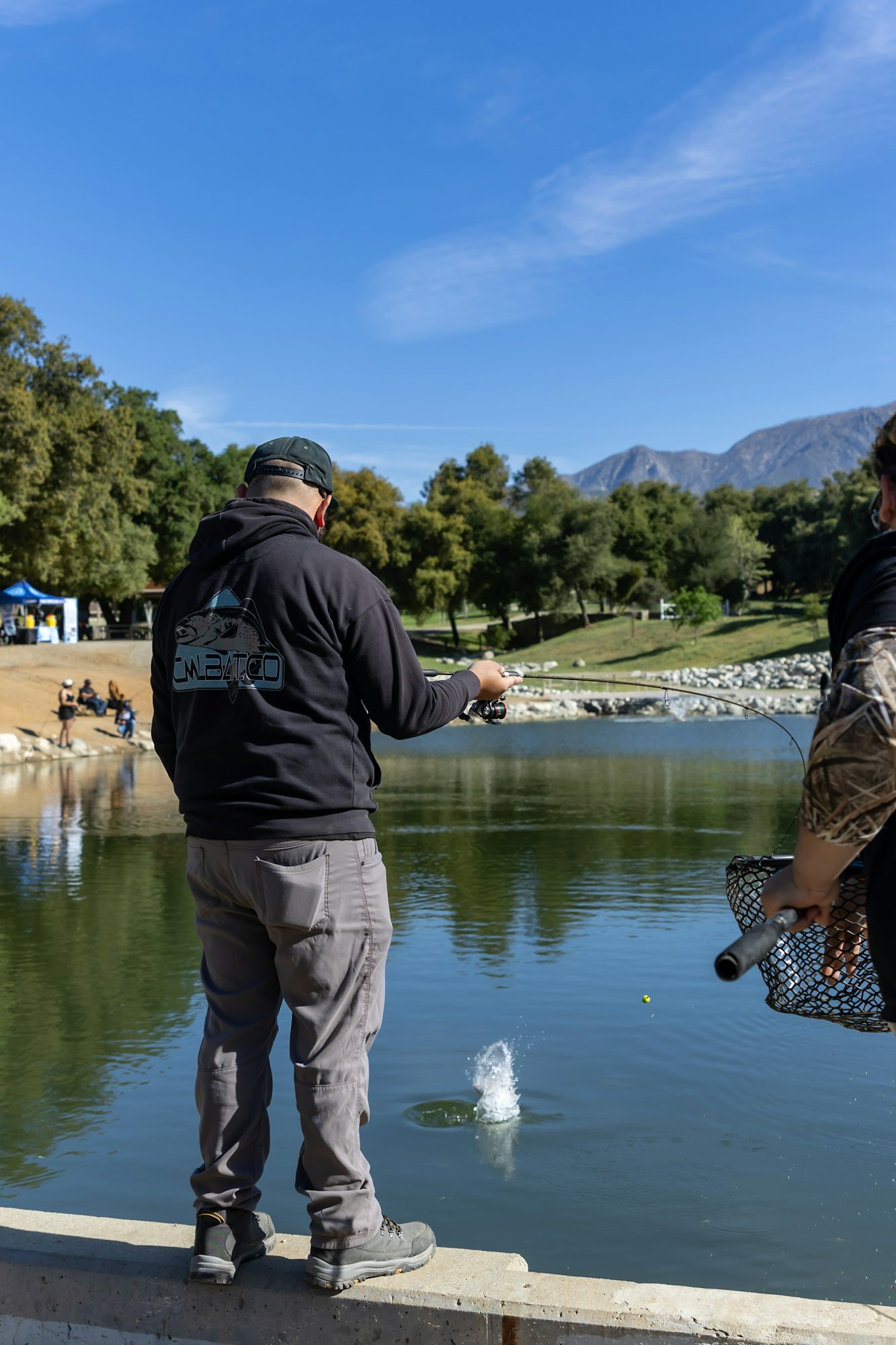 People fishing by a pond, one holding a rod and another with a net, surrounded by trees and mountains.