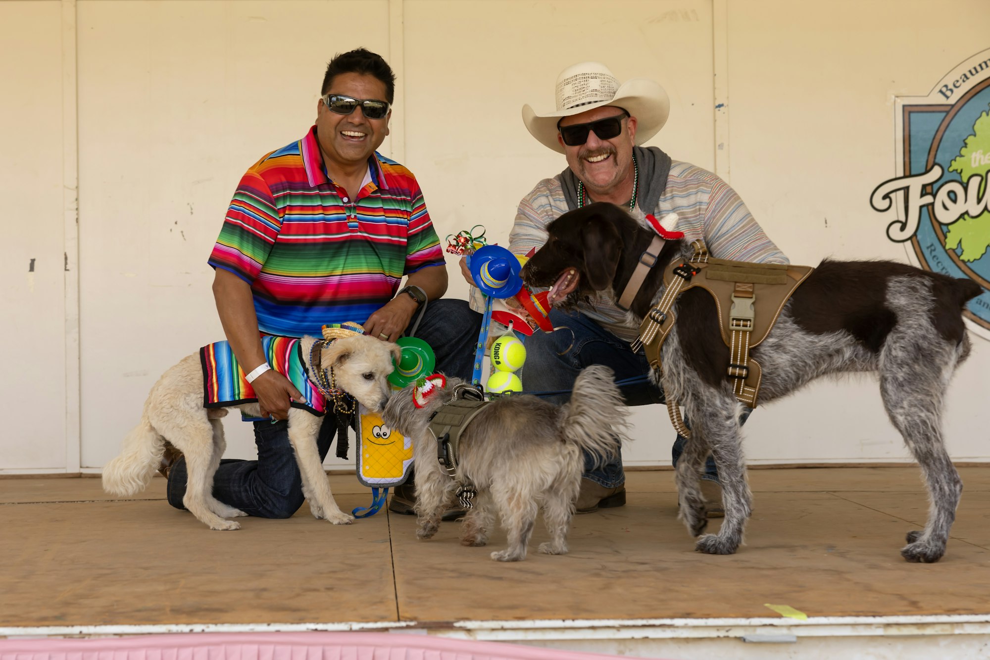 Two smiling men and three dogs in colorful outfits posing together on a stage.