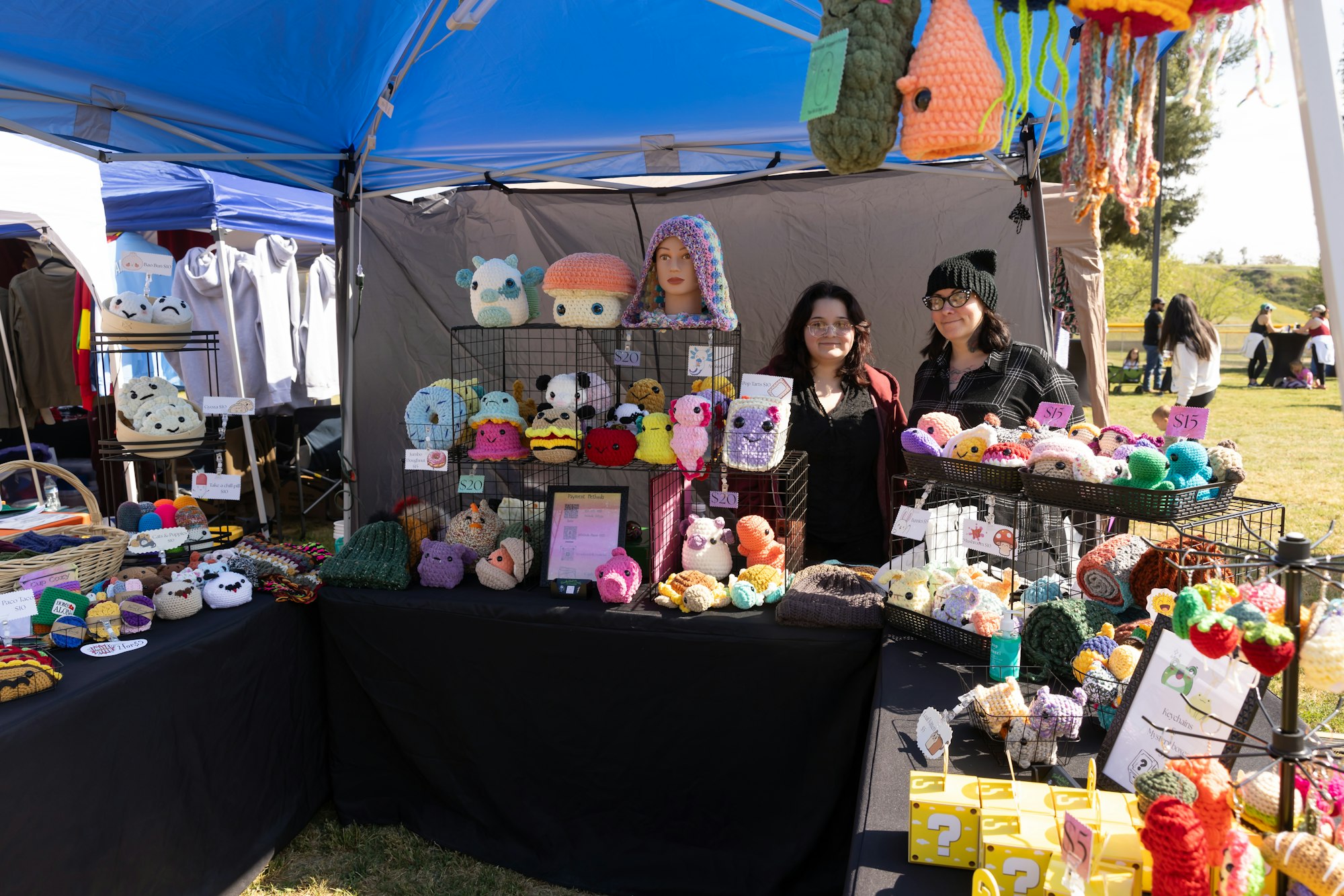 Outdoor craft market booth displaying crochet items and plush toys, operated by two people under a blue canopy.