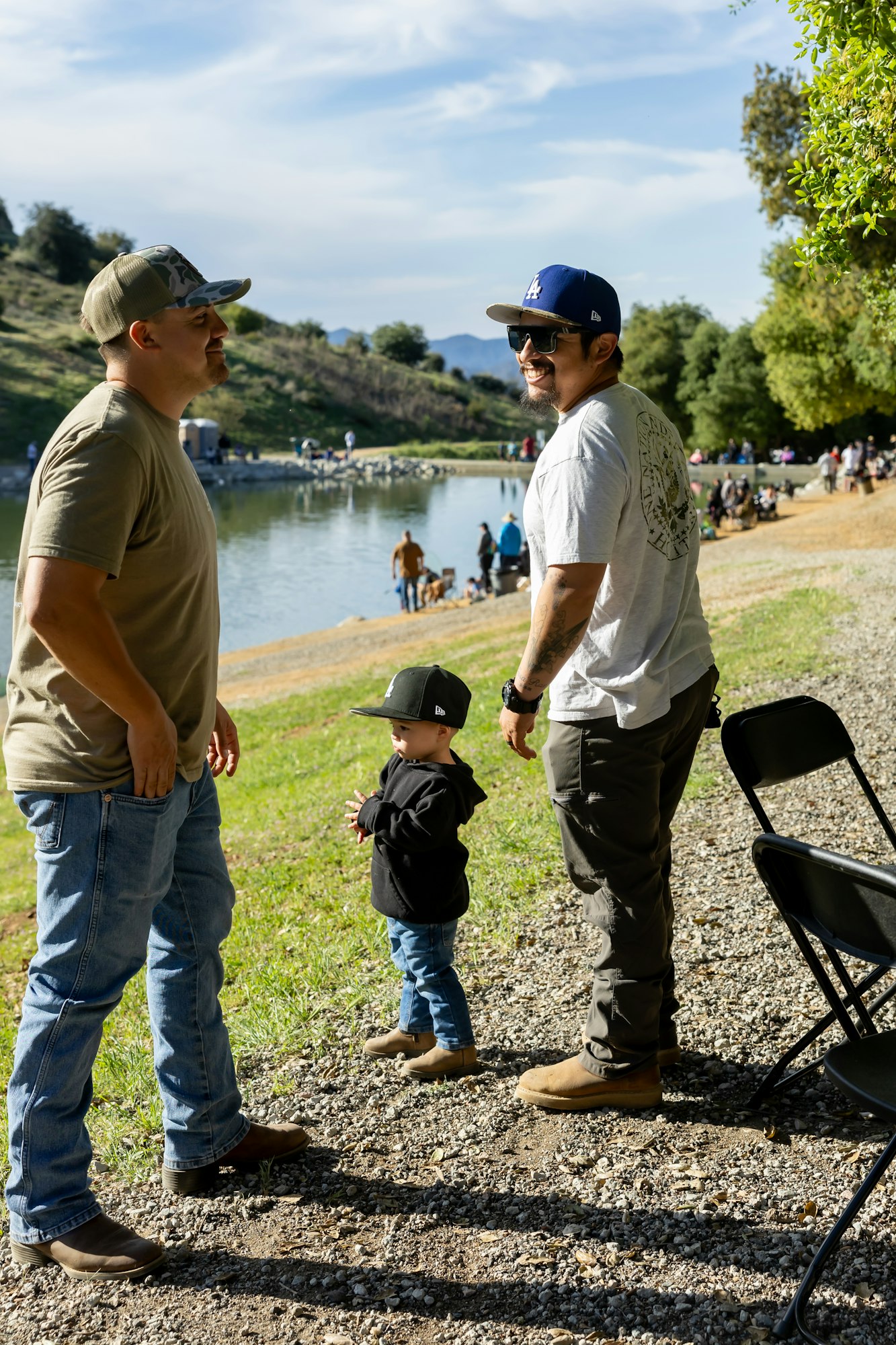 Two adults and a child in hats smiling by a lake with people and trees in the background.