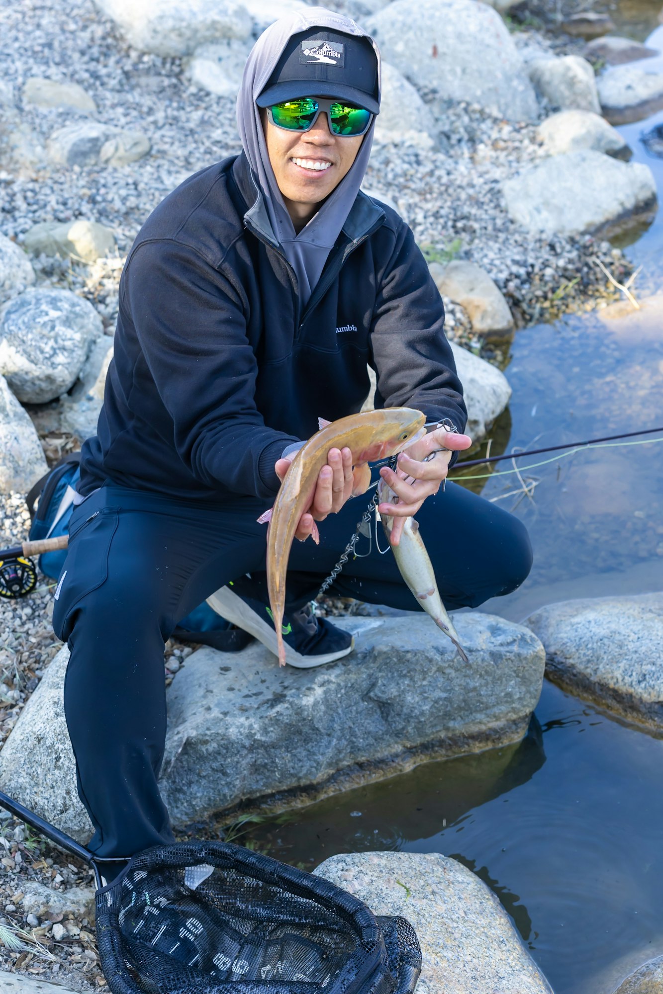 A person wearing sunglasses and a hoodie is holding a fish by a rocky stream, with a net on the ground.