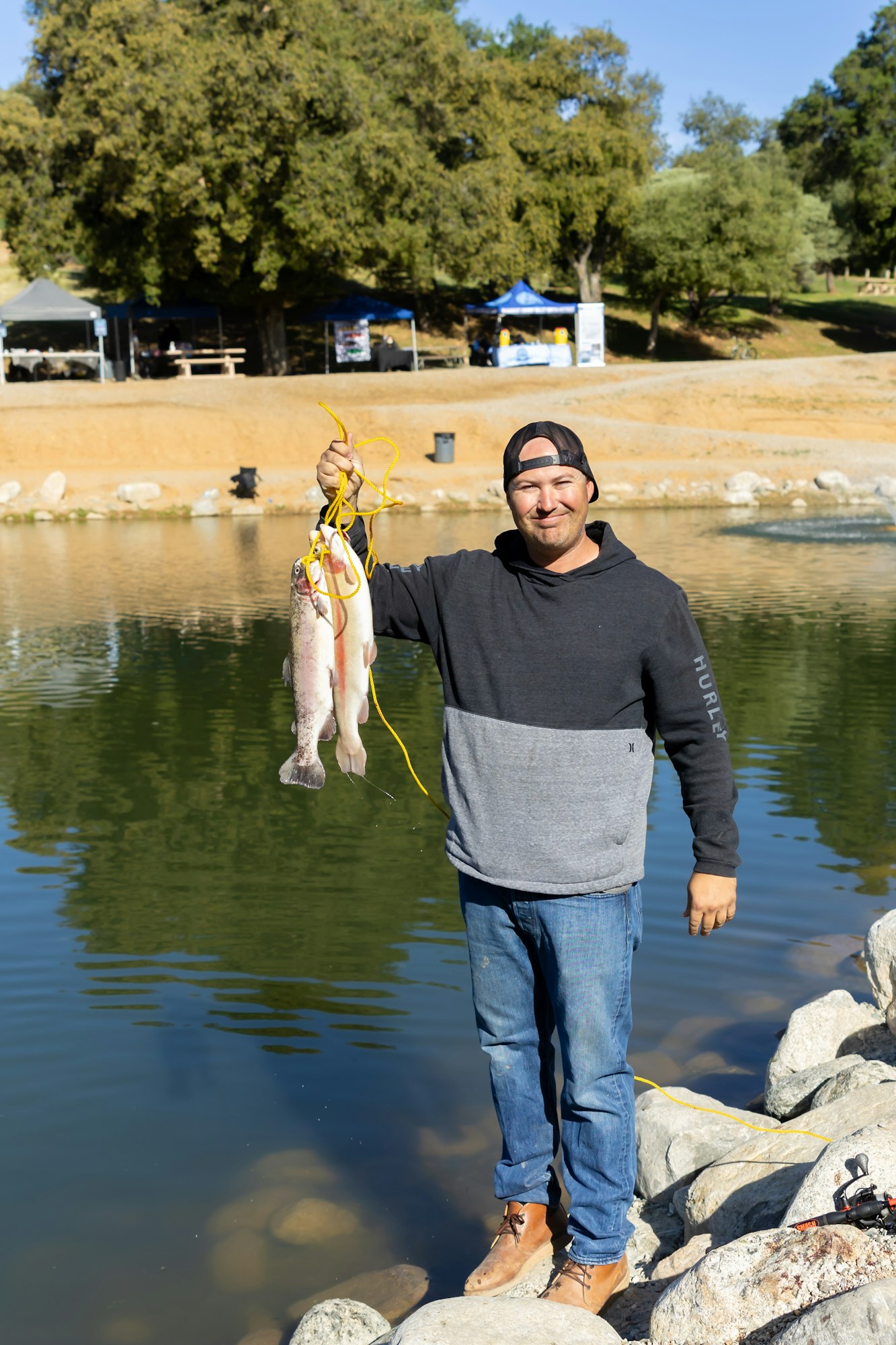 A person stands on rocks by a pond, holding a stringer with two fish, wearing a cap and casual clothes. Trees and tents are in the background.