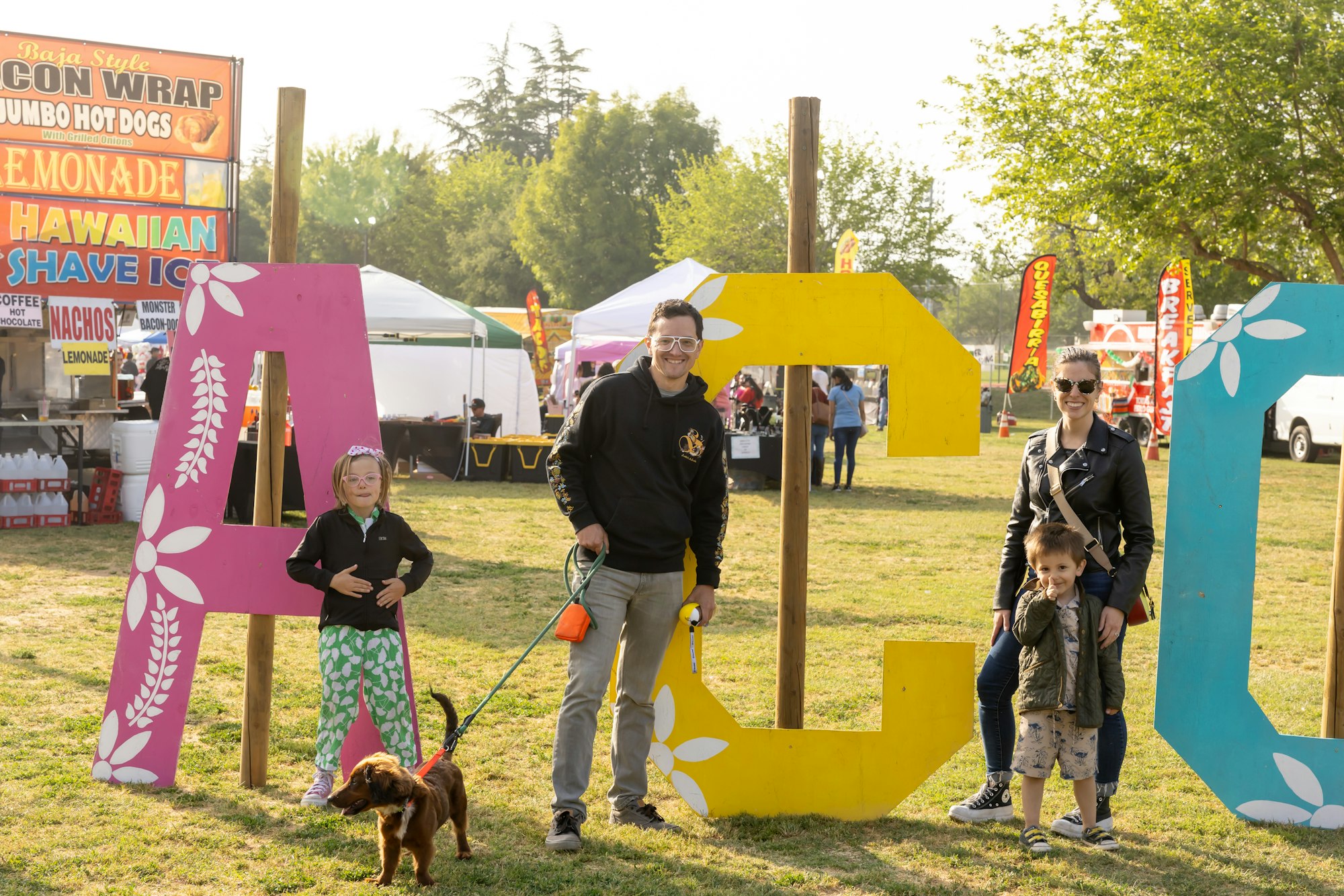 A family with a dog poses in front of large A, C, O letters at an outdoor event with food stalls in the background.