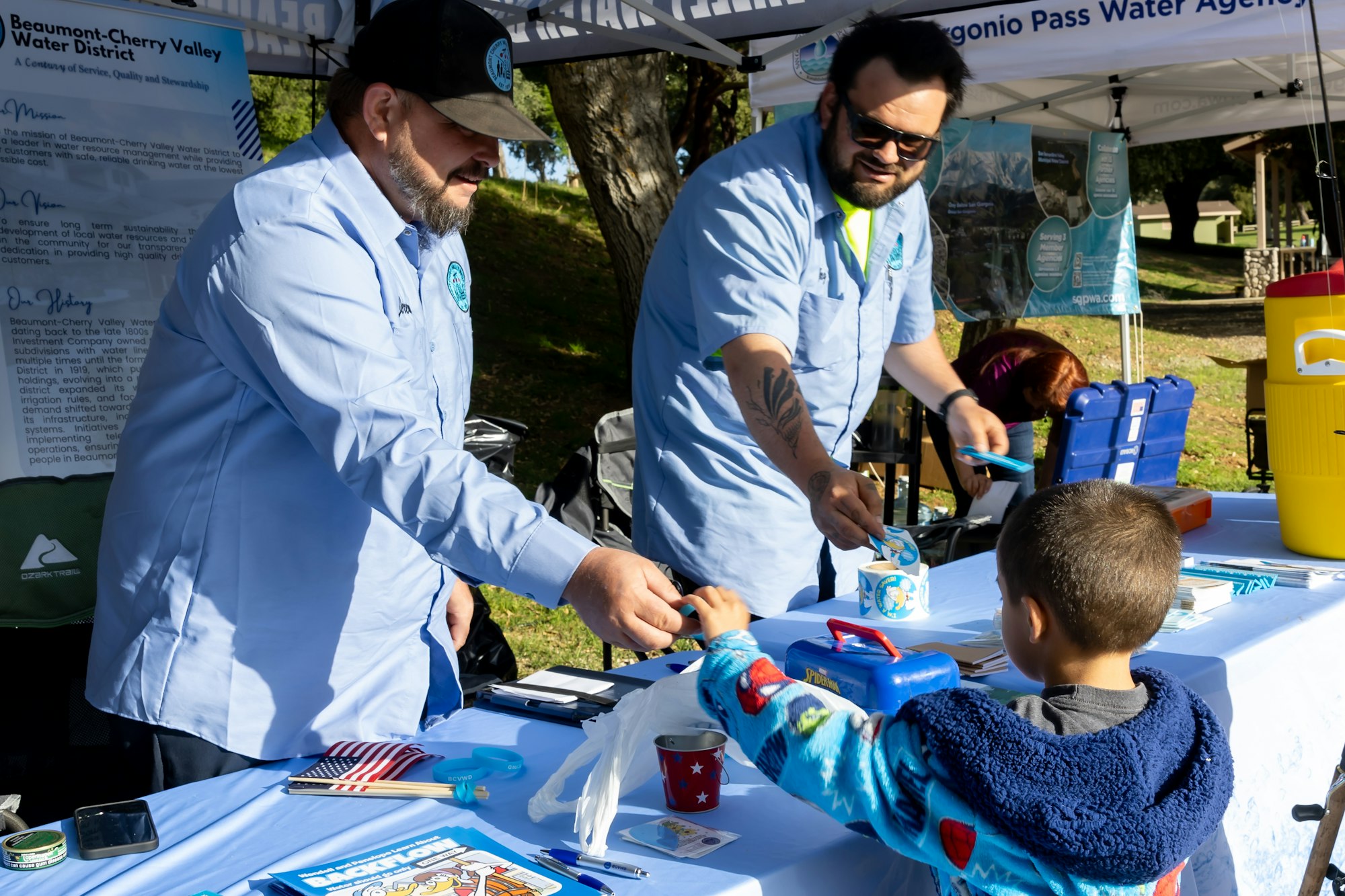 Two men at a booth hand out items to a child, with a banner for Beaumont-Cherry Valley Water District in the background.