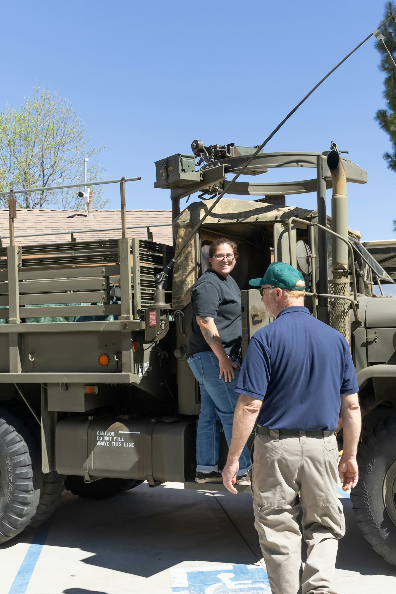 A woman is getting into a military vehicle while a man in a cap approaches. It's a sunny day with clear blue skies.