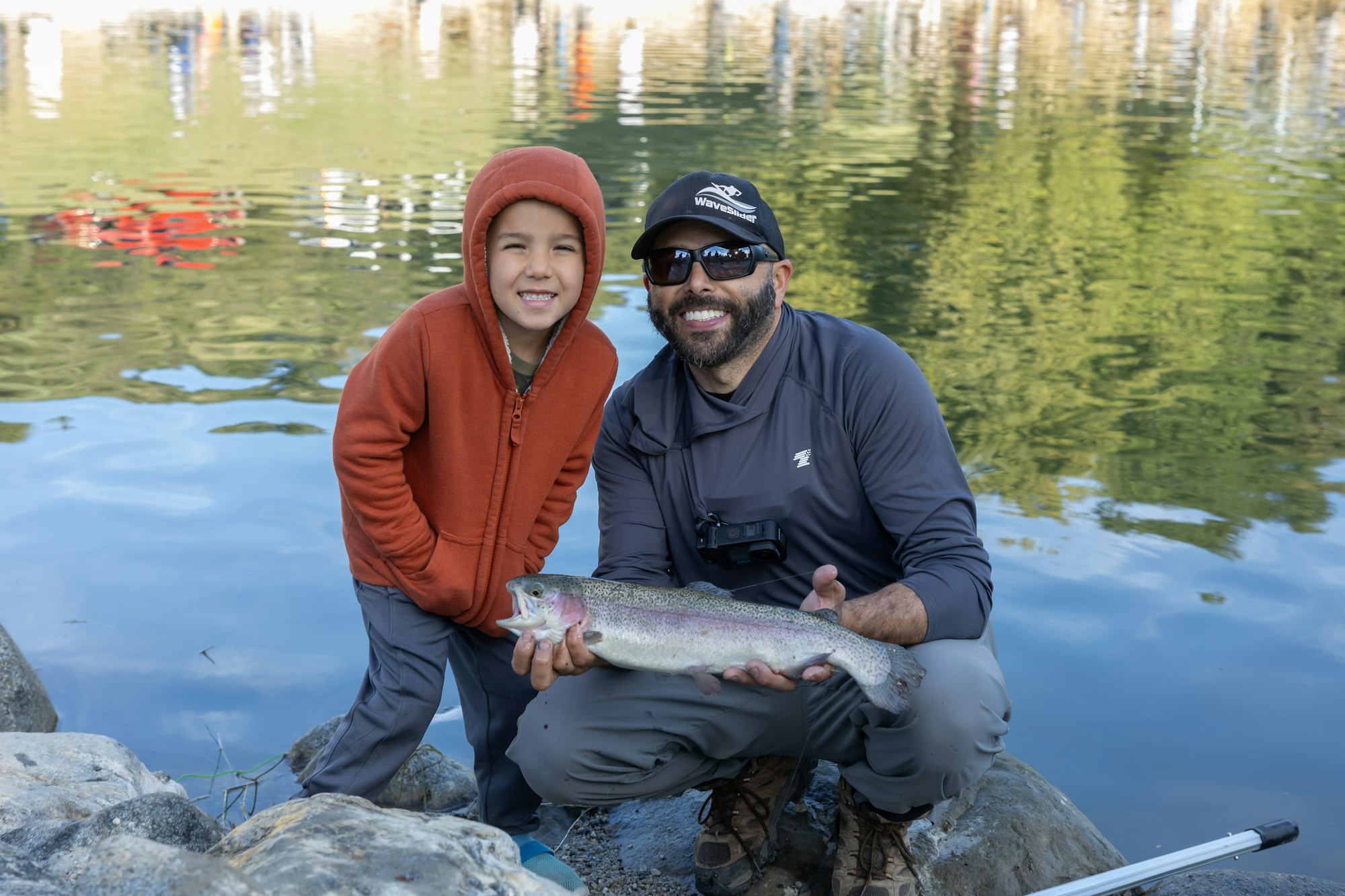 Two people smiling by a lake, one holding a caught fish.