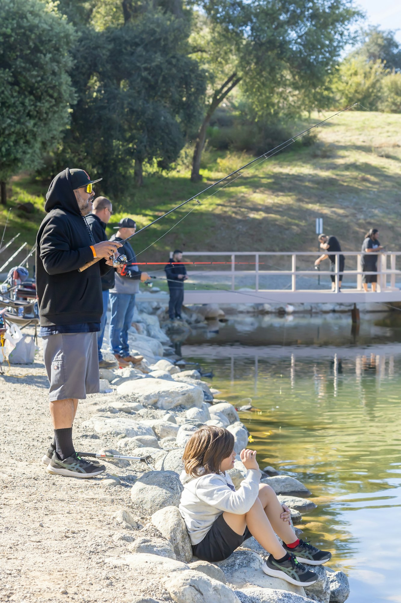People fishing by a lake, with one child sitting on rocks.