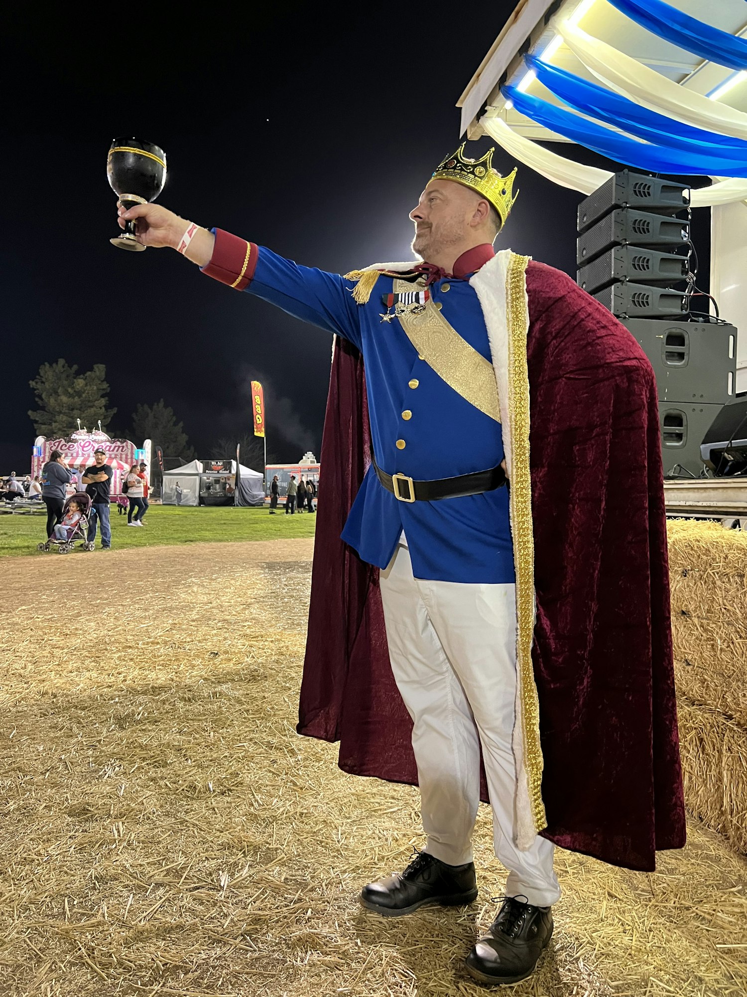 A person in royal costume with a crown and cape holds a goblet, standing on a straw surface outdoors at night.